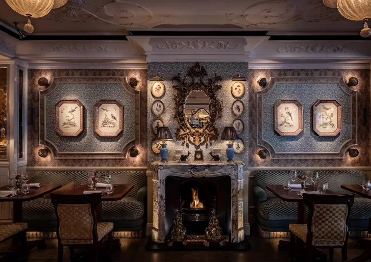 Elegant restaurant interior with ornate wallpaper, framed bird illustrations, and a white marble fireplace topped with a decorative mirror, candles, and blue vases. Cozy tables and plush seating line both sides of the room.