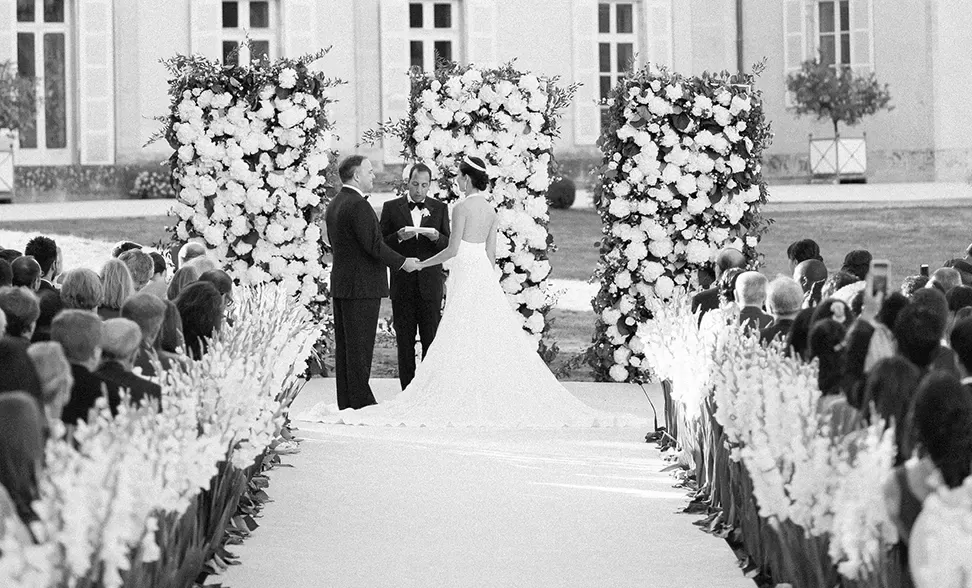 A black and white image of a wedding ceremony. A couple stands before an officiant, surrounded by tall floral arrangements. Guests are seated on either side of an aisle lined with flowers, and a building is visible in the background.