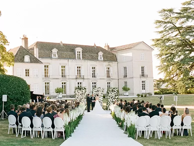 A beautiful outdoor wedding ceremony is set up in front of a grand, light-colored historic mansion. Rows of white chairs line a white aisle adorned with floral arrangements. Guests are seated, and the couple stands facing the officiant, surrounded by greenery.