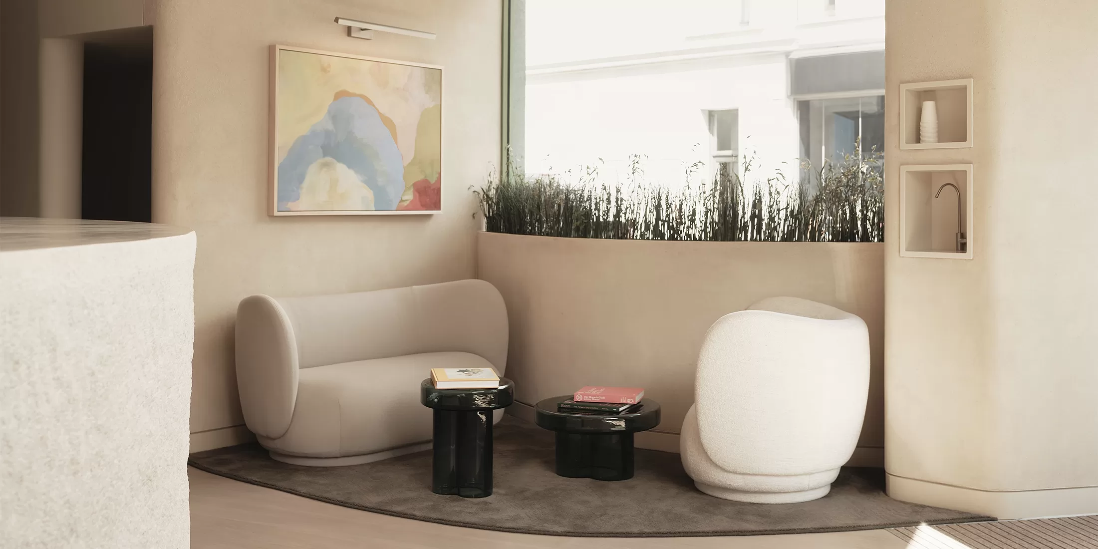 A modern living room with a minimalist design featuring a curved sofa and chair. Two small black tables display books. A large abstract painting hangs on the wall, and a tall window lets in natural light, highlighting a planter with greenery.