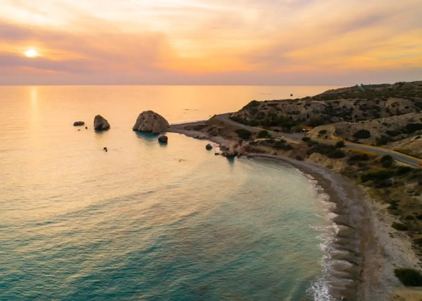 Aerial view of a rocky coastline at sunset, with calm turquoise water, a winding coastal road, and several large rocks emerging from the sea near the shore under a colorful sky.