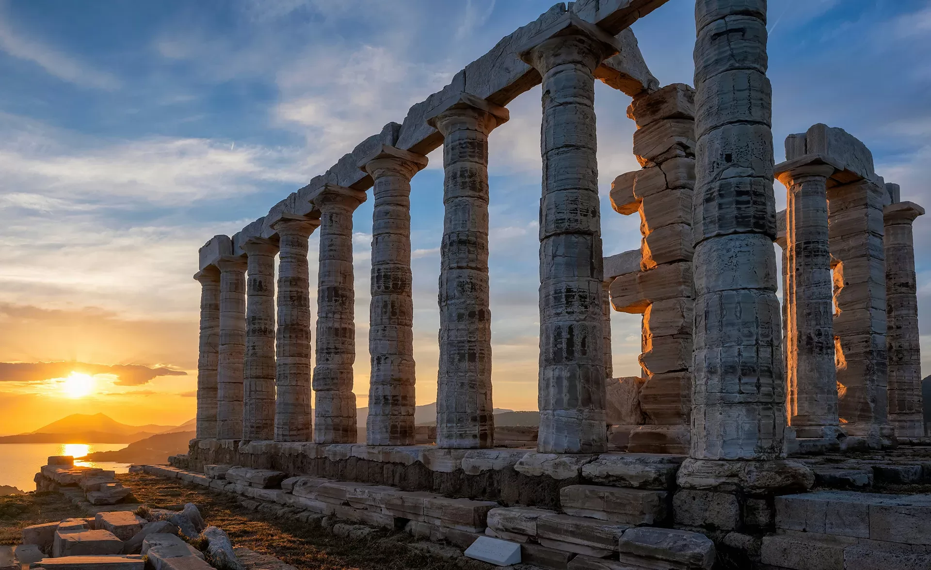 The image shows the ancient Temple of Poseidon at Cape Sounion, Greece, with tall stone columns. A vibrant sunset in the background casts a warm glow, and the sky is partly cloudy with hints of orange and blue.