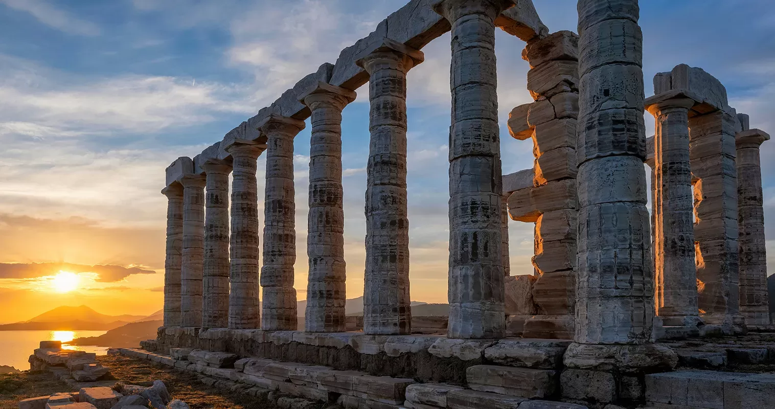Ancient stone columns of the Temple of Poseidon at Cape Sounion, Greece, stand tall against the vibrant sunset sky. The sun is near the horizon, casting a warm glow on the ruins and creating a dramatic, picturesque scene.