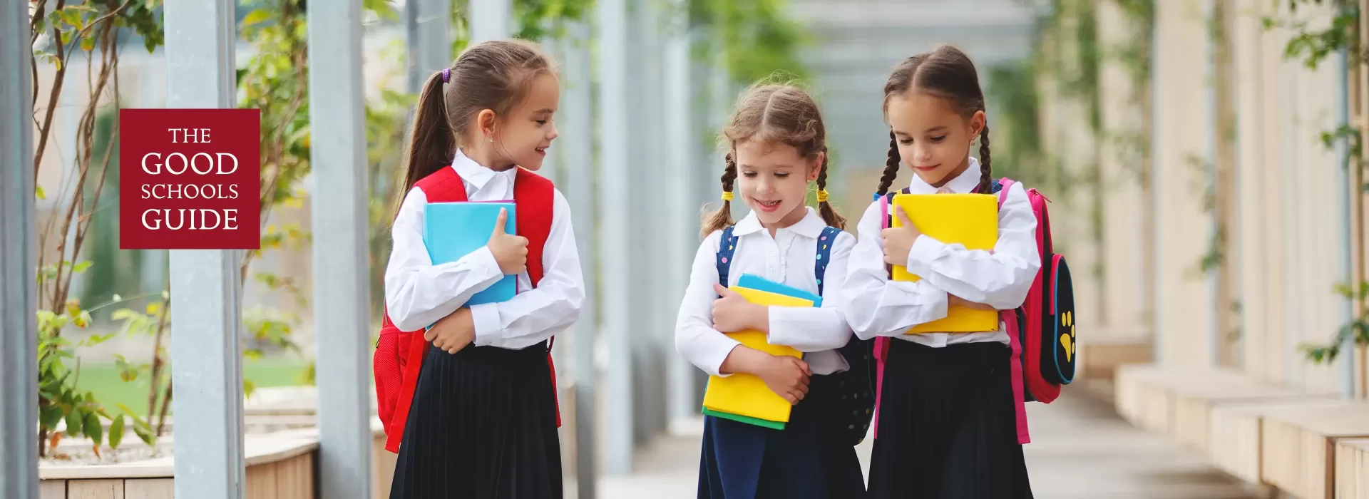 Three young schoolgirls in uniform walk outdoors, smiling and carrying colorful books and backpacks. A sign on the left reads The Good Schools Guide. The background features greenery and columns.