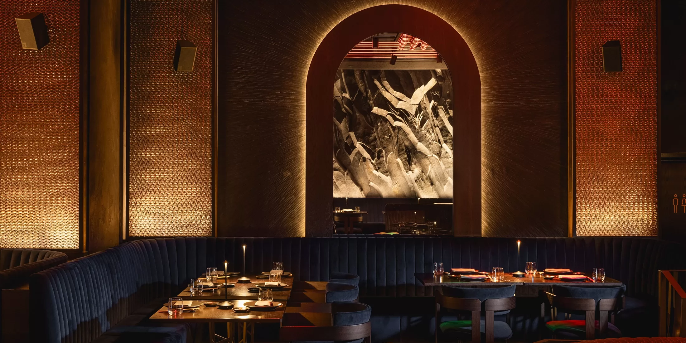 Dimly lit, elegant restaurant with dark velvet seating, round tables set for dining, and a dramatic arched opening framing a textured black-and-white wall art in the background, creating a sophisticated ambiance.