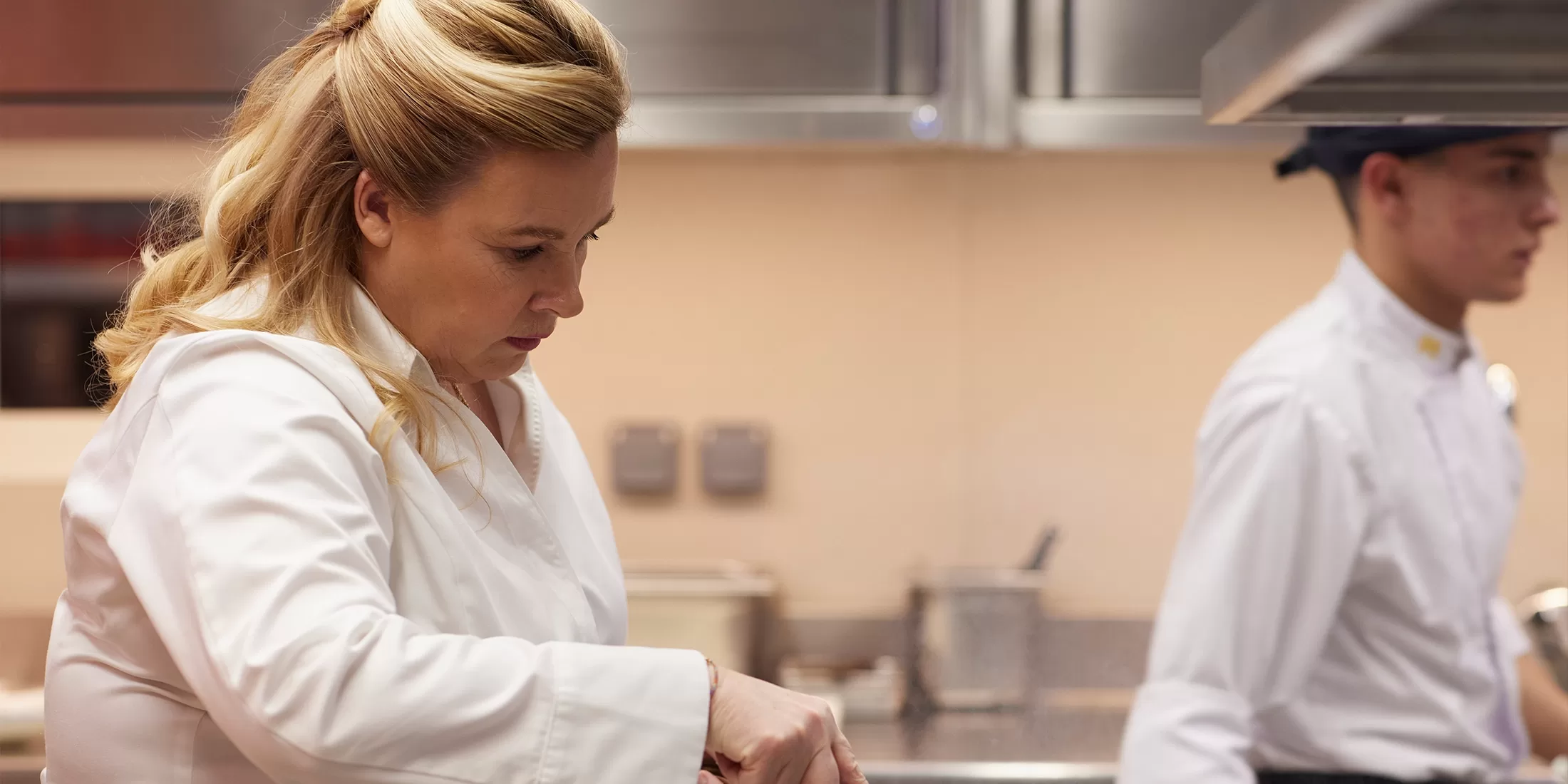 A woman in a white chefs coat prepares food in a professional kitchen, while a man in a chefs uniform stands blurred in the background.