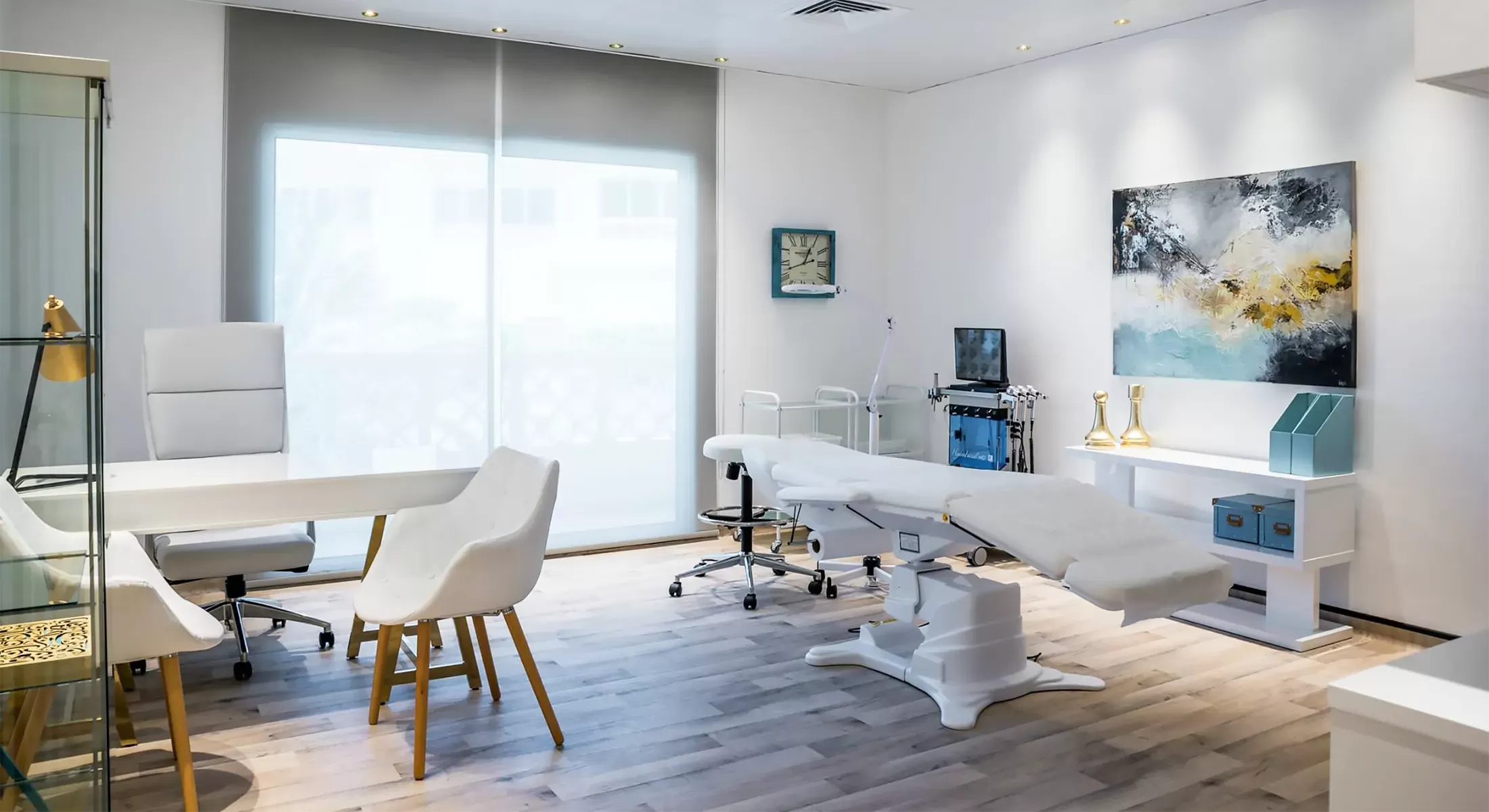 Beautician room in Dubai featuring a white treatment bed, white table and chairs, wooden flooring at painting on the wall.