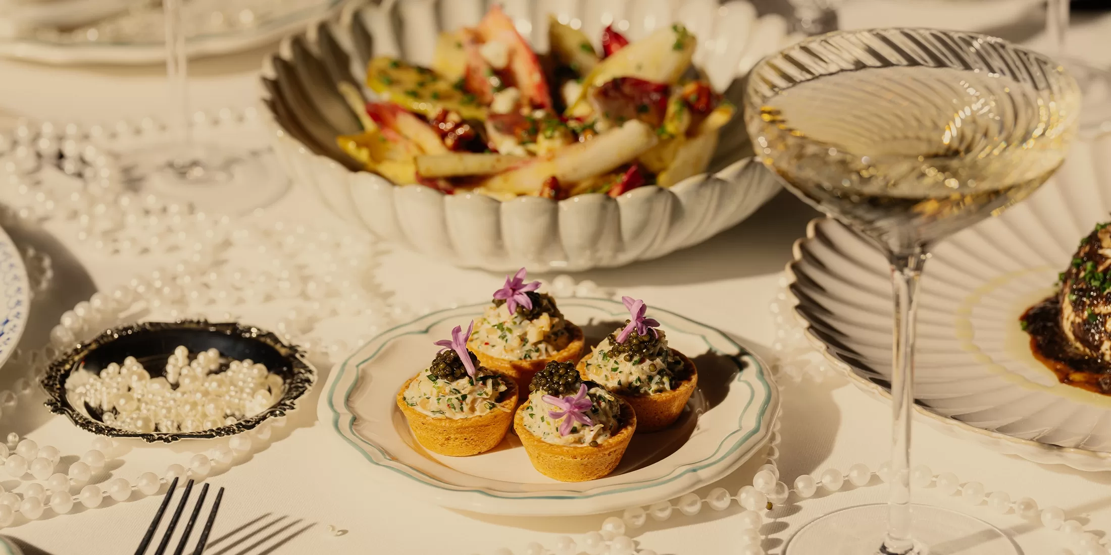 A plate of elegant bite-sized appetizers garnished with purple flowers is set on a table with a salad bowl, glass of white wine, black caviar, and decorative pearl strands in the background.