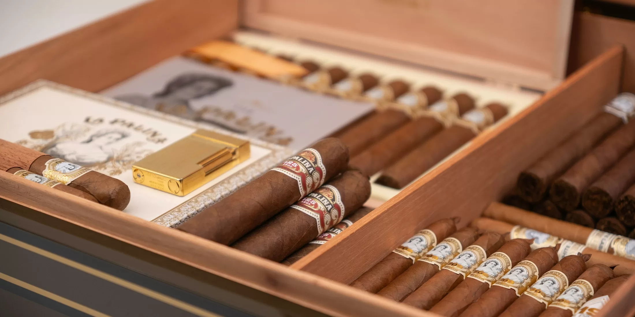 An open wooden humidor box filled with neatly arranged cigars, a gold cigar cutter, and a booklet featuring a portrait, all organized in separate compartments.