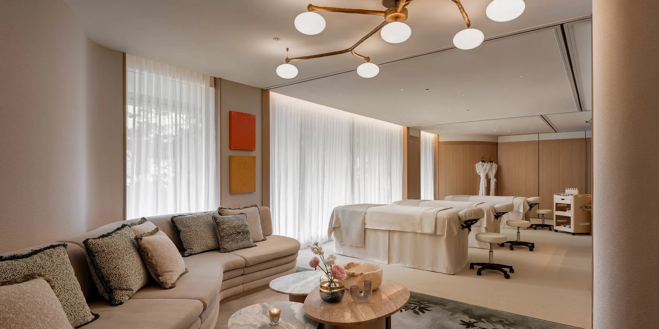A luxurious spa room with two massage tables, modern beige furnishings, a plush sofa with cushions, soft lighting, and a contemporary chandelier. Sheer curtains cover the windows, creating a calm and inviting atmosphere.