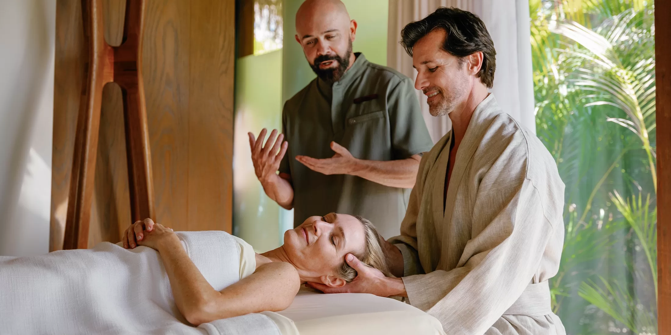 A woman lies on a massage table with her eyes closed, covered by a towel, while a man in a robe gently supports her head. Another man stands nearby, gesturing as if explaining or guiding the process.