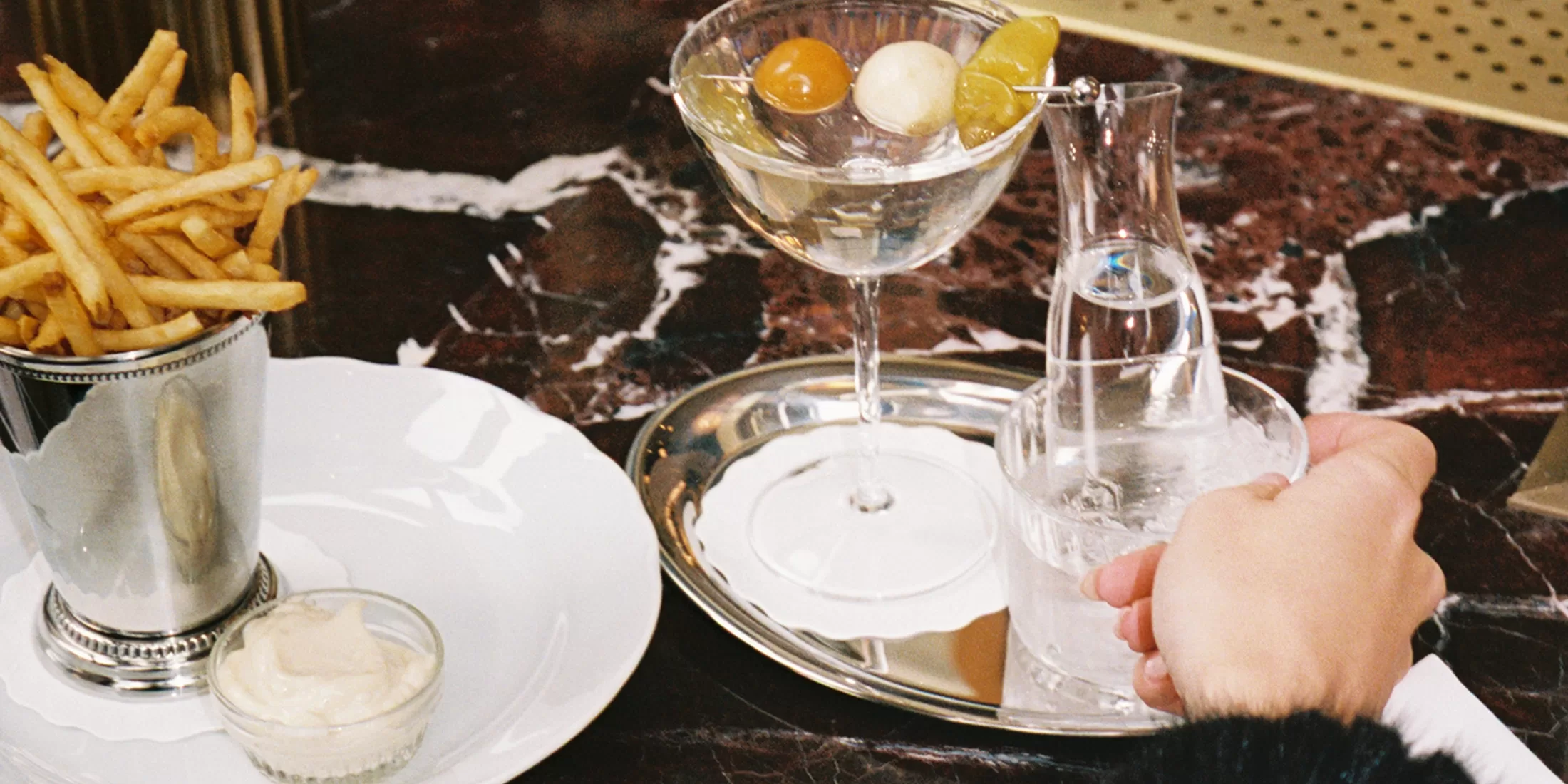 A hand pours water from a small carafe into a glass on a silver tray beside a cocktail with olives and garnish, french fries in a metal cup, and a bowl of dipping sauce on a marble table.