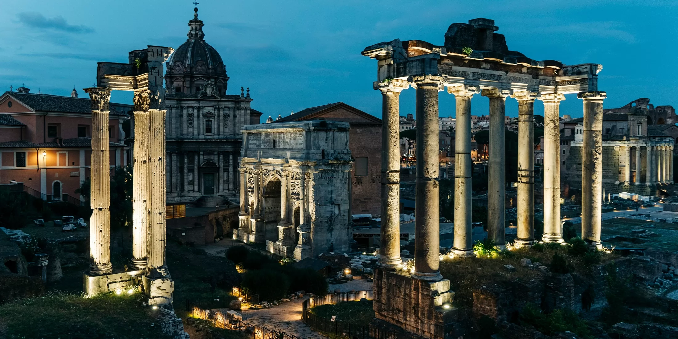 Ancient Roman ruins with tall stone columns and arches illuminated at dusk, with a domed church and historic buildings in the background under a deep blue sky.