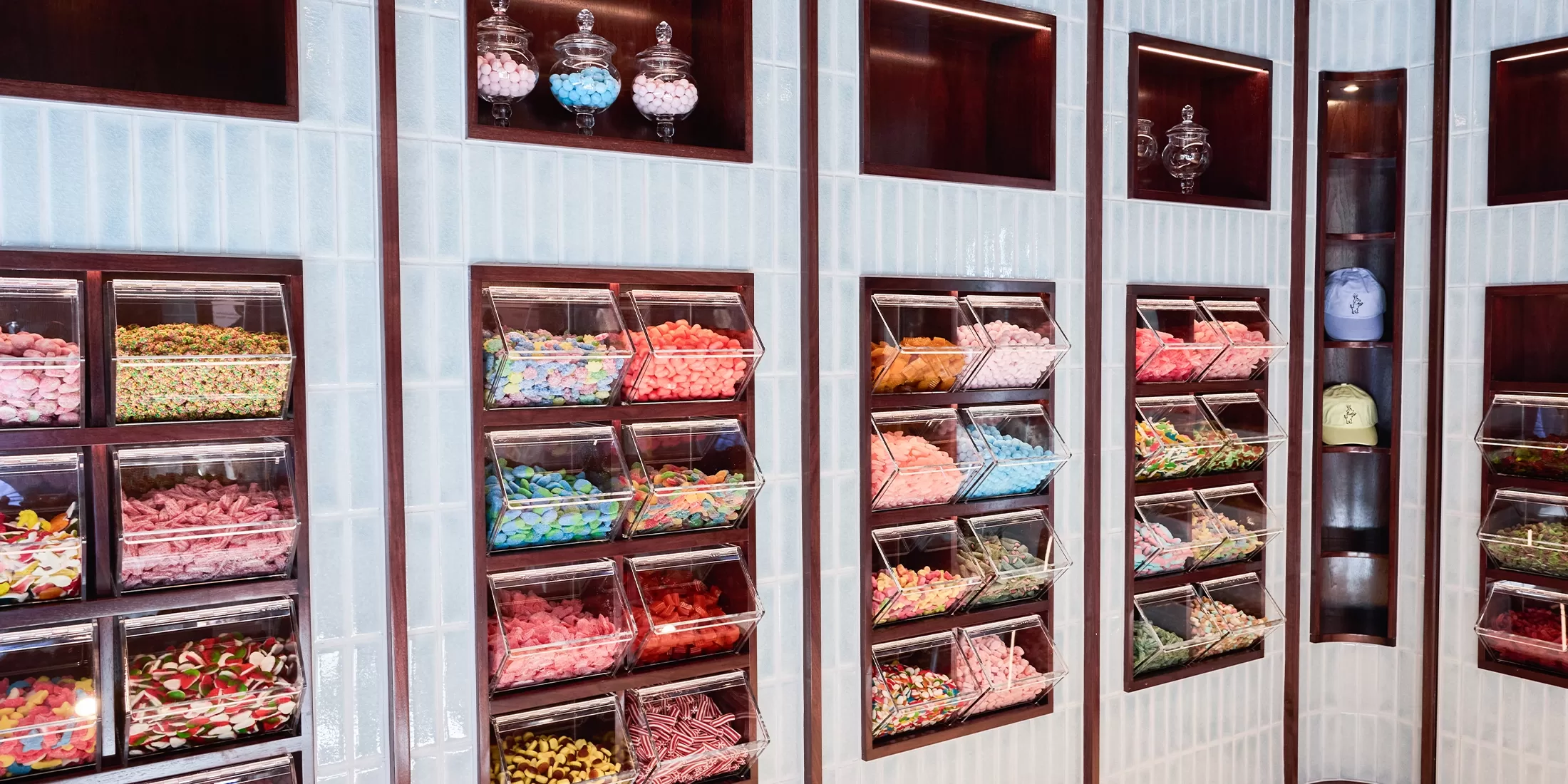 A wall display of clear bins filled with assorted colorful candies, including gummies and licorice, in a modern candy shop. Glass jars with more sweets are on upper shelves.