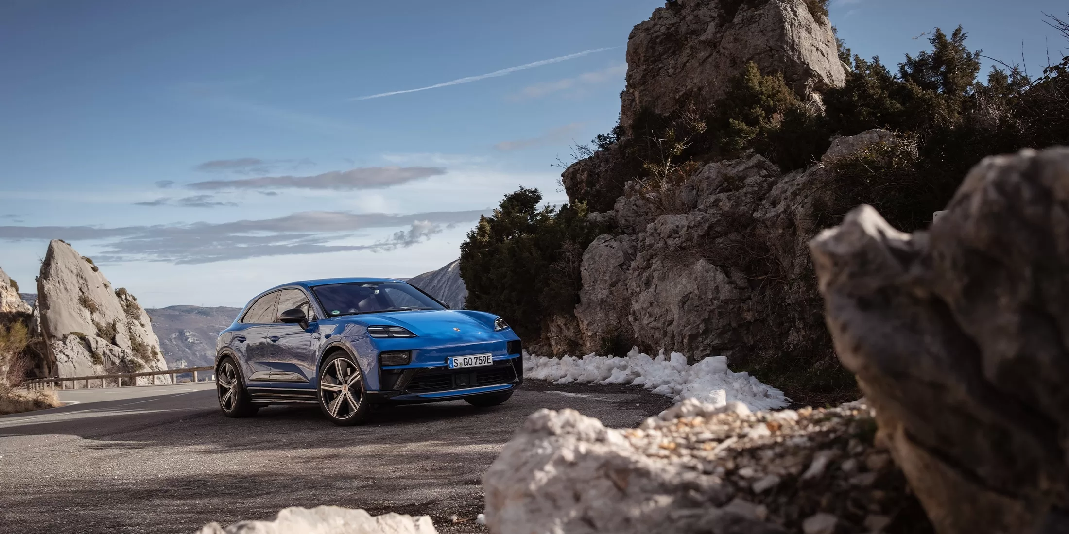 A blue SUV driving on a mountainous road, surrounded by rocky cliffs and patches of snow, under a clear blue sky.