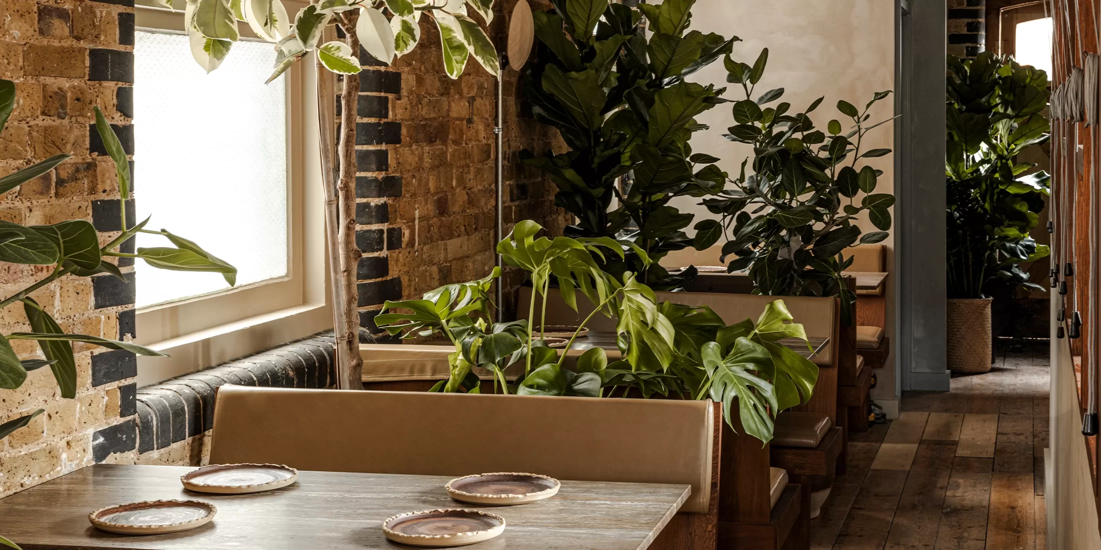 A cozy restaurant interior with wooden tables set with plates, tan cushioned booths, exposed brick walls, large windows, and lush green potted plants creating a warm, inviting atmosphere.