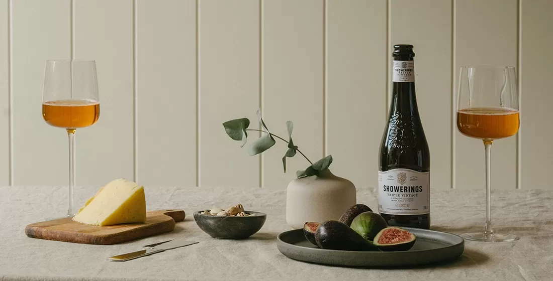 Table set with plates and boards of cheese, fruit, a vase with flower and Showering's cider with two glasses