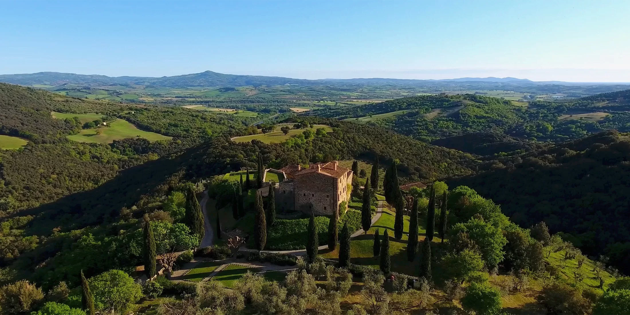 Aerial view of a rustic villa surrounded by tall cypress trees on a lush, green hillside. In the background, gentle rolling hills and distant mountains under a clear blue sky.