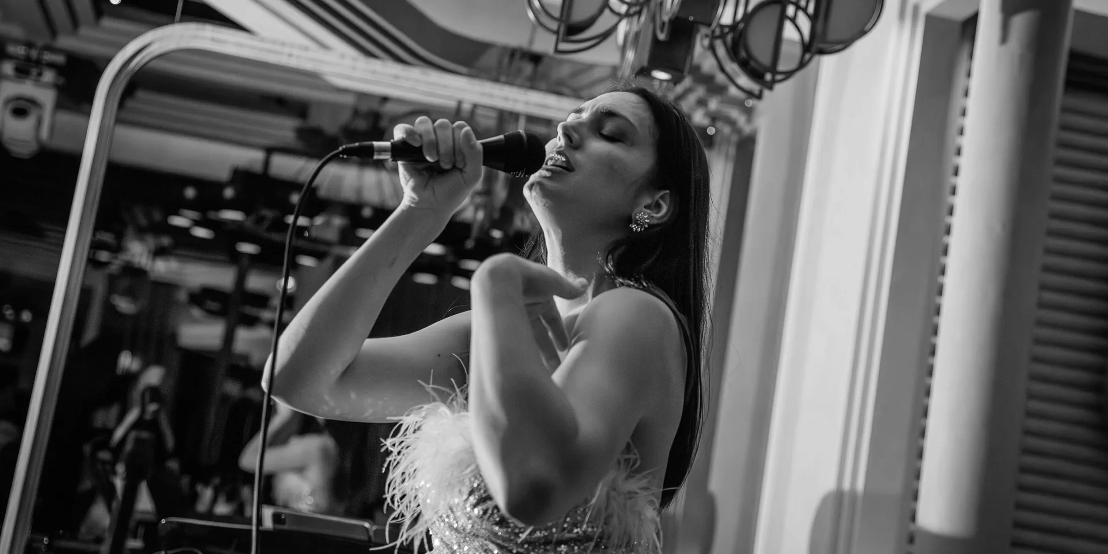 Black and white photo of a woman singing passionately into a microphone. She wears a dress adorned with feathers and sequins. Her eyes are closed, and she holds the microphone with one hand, gesturing with the other.