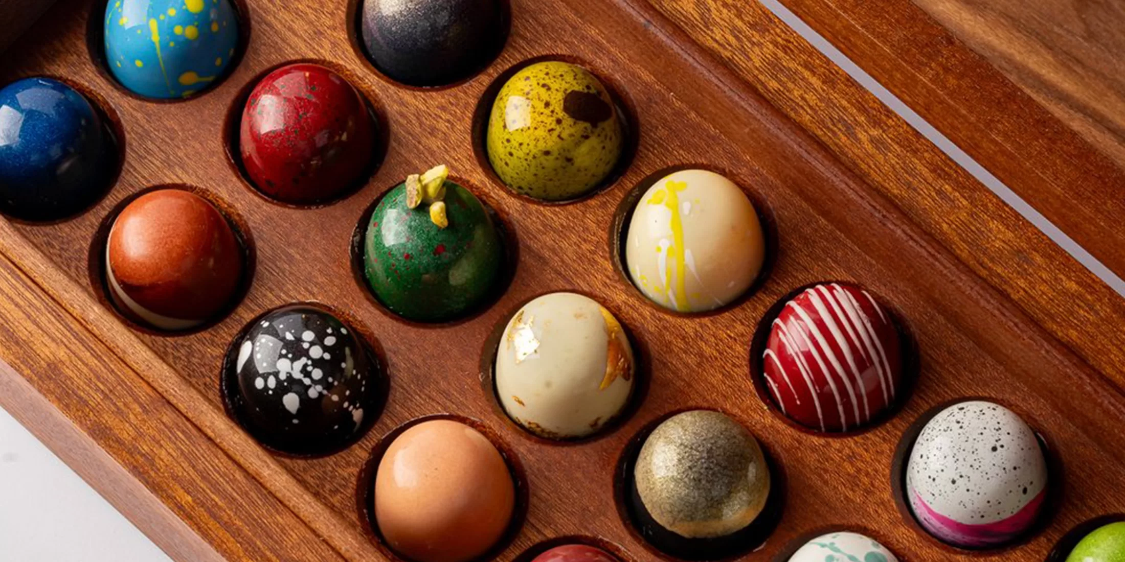 A wooden box holds colorful, dome-shaped chocolates with various patterns, including speckles, swirls, and stripes, arranged neatly in individual compartments by Adam Handling Chocolate Shop.