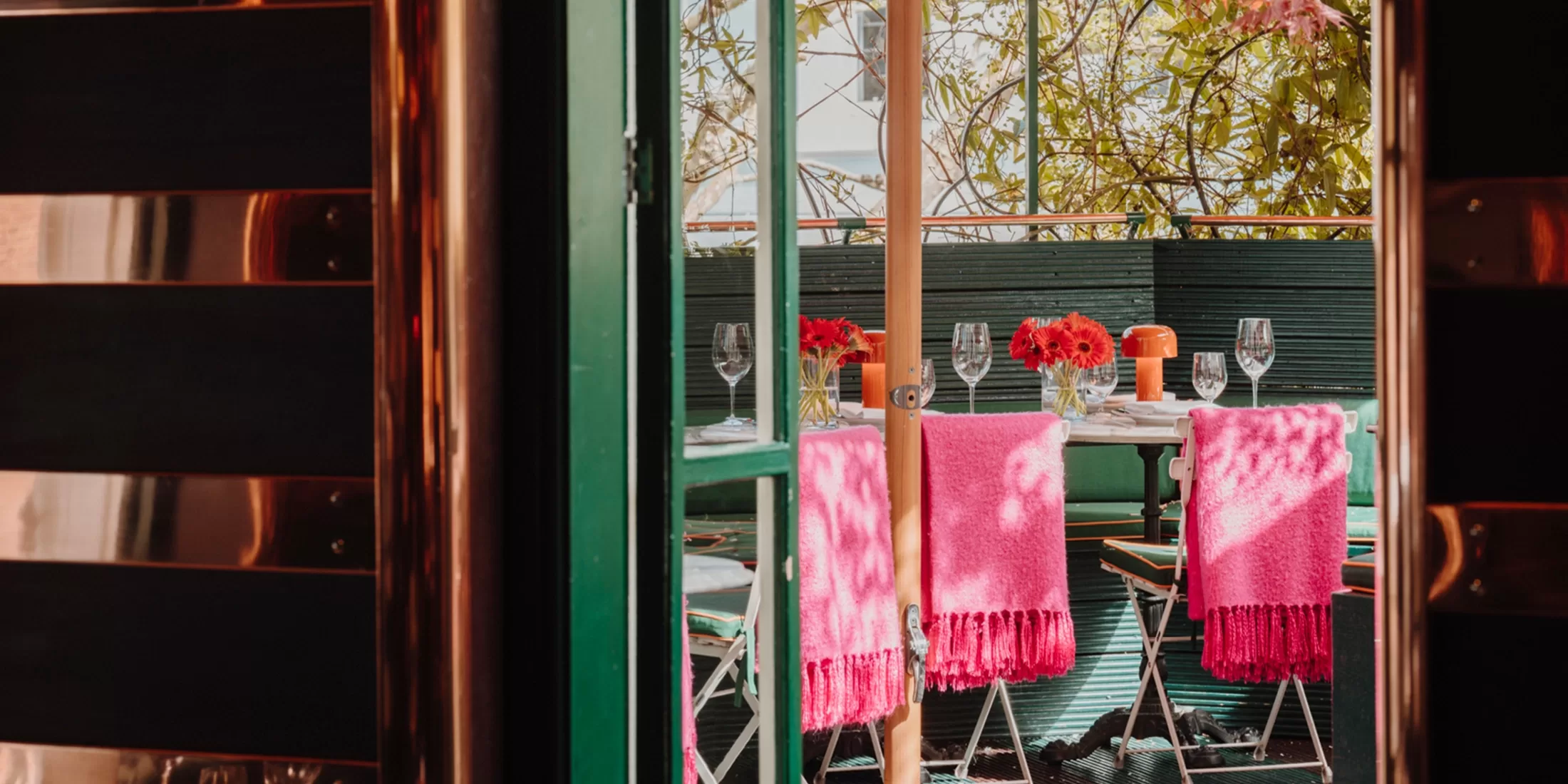 Outdoor dining area with a table set for four. Bright pink blankets cover two backrests. Red flowers in vases, wine glasses, and place settings adorn the table. Green walls and leafy branches add a cozy, natural ambiance.