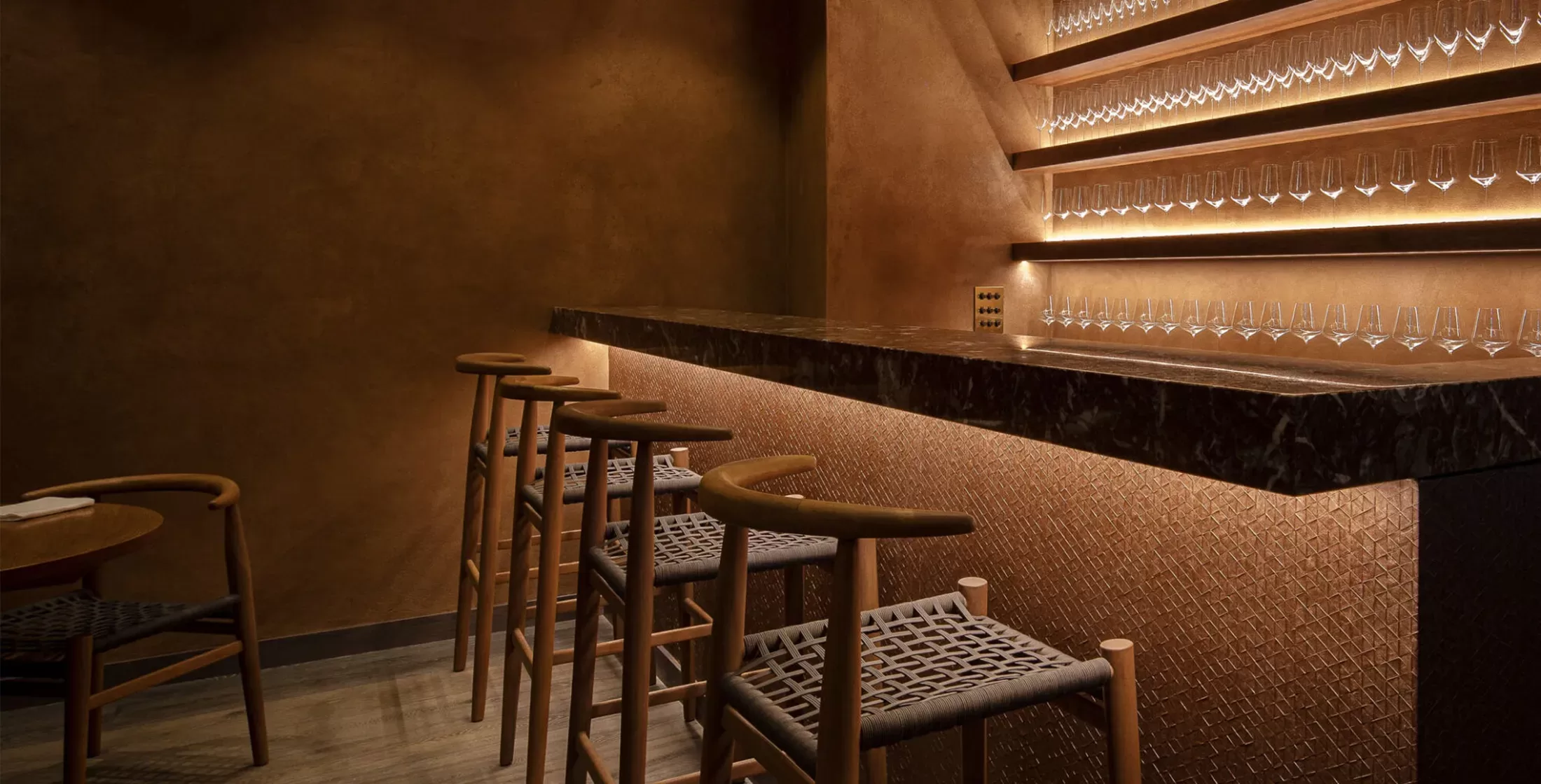 A dimly lit bar with a sleek black marble counter and wooden stools. The wall behind the counter features rows of neatly arranged glasses illuminated by warm lighting, creating an inviting and elegant atmosphere.