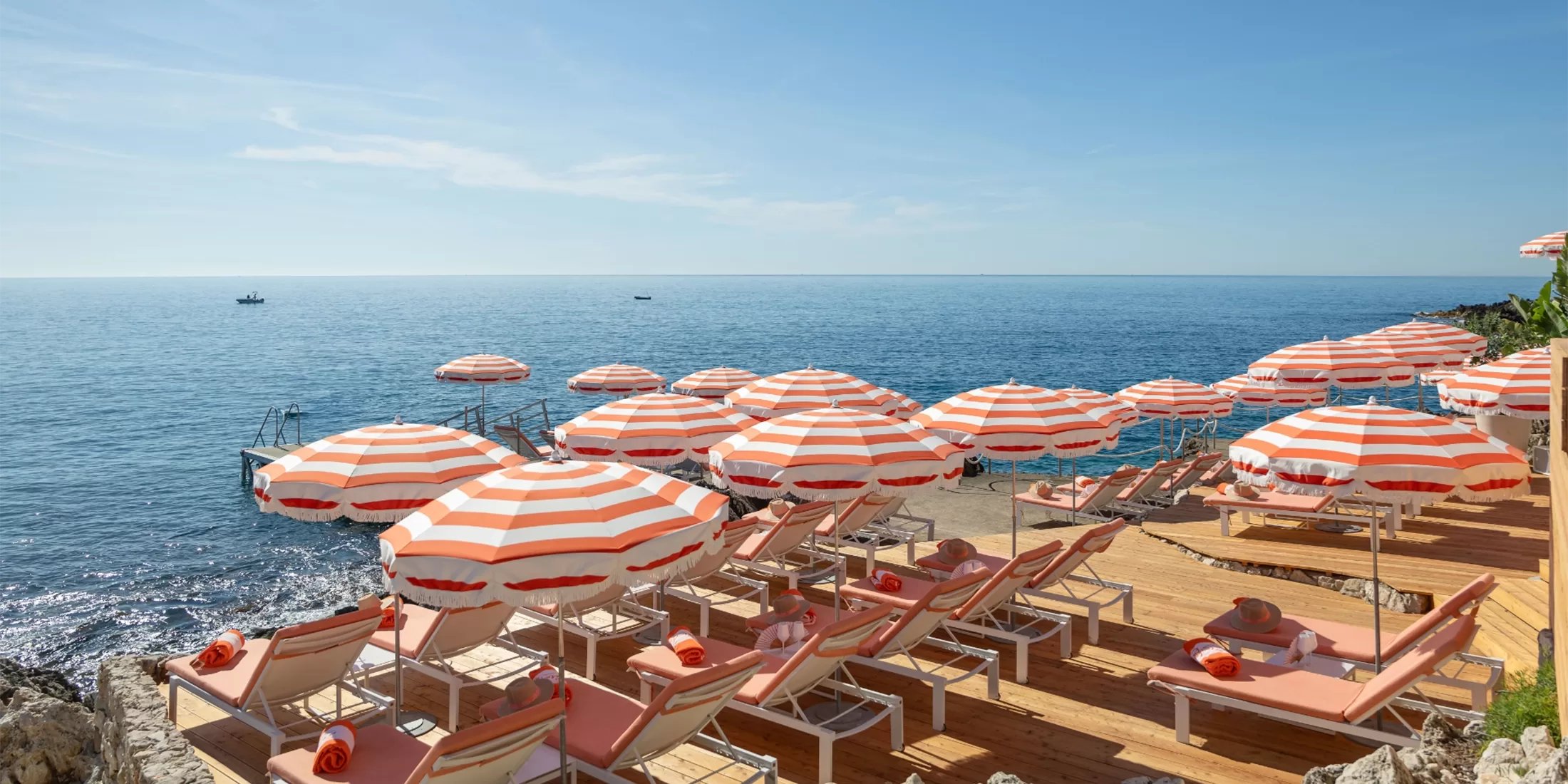 Rows of orange-and-white striped umbrellas and sun loungers face a calm blue sea on a sunny day, creating a relaxing, vibrant beachside scene.