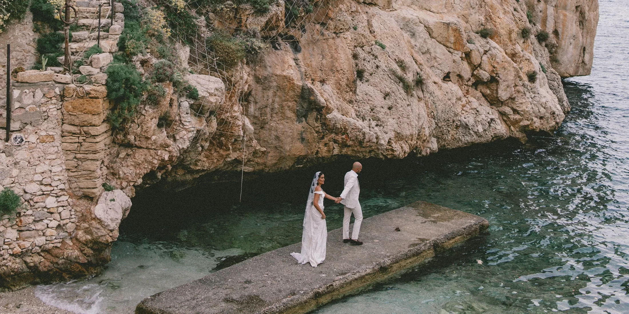 A bride and groom stand on a concrete platform by clear, rocky waters, holding hands and facing each other, with large stone cliffs and greenery in the background.