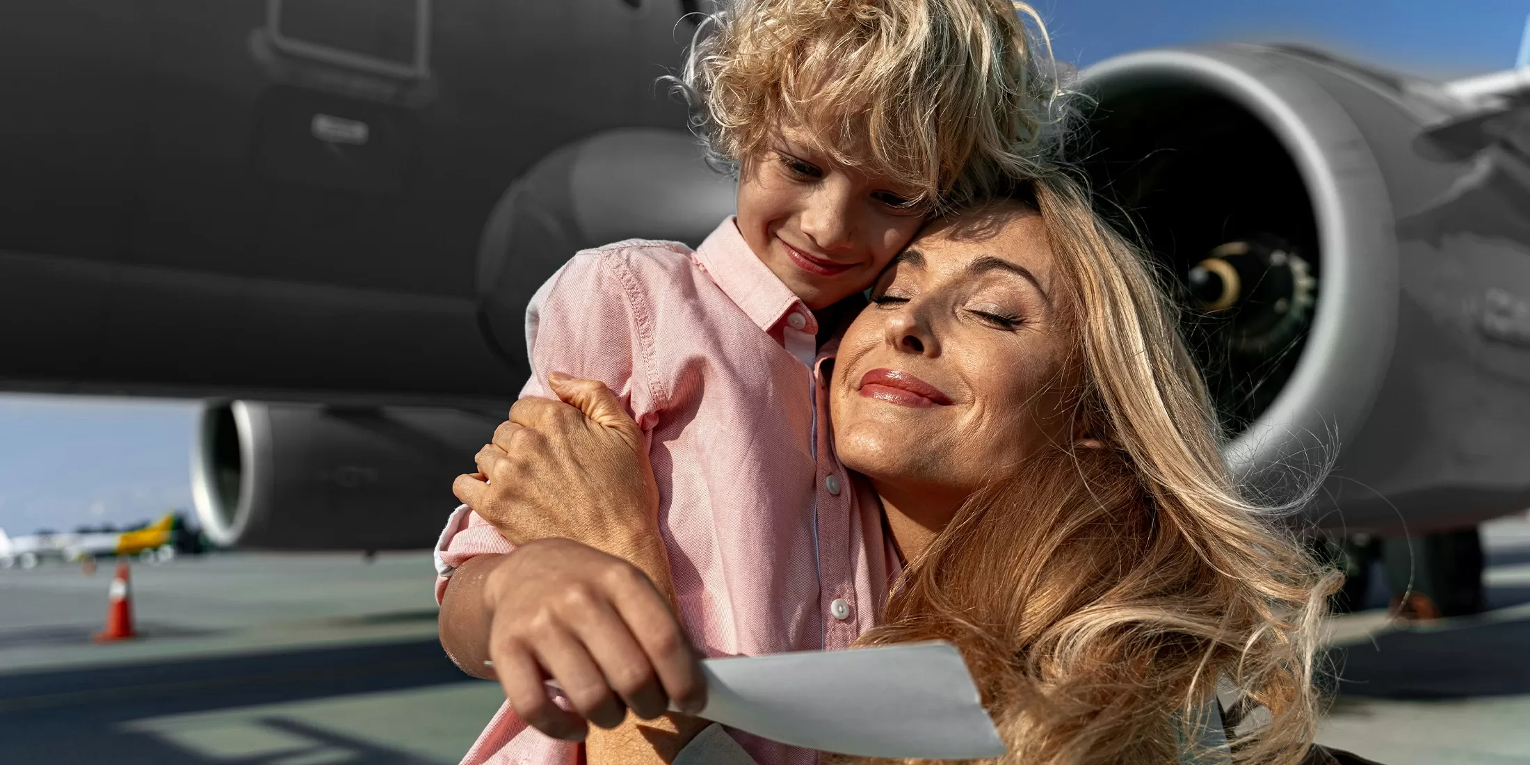 A smiling woman hugs a young boy tightly in front of an airplane on a sunny day. She holds a piece of paper, possibly a ticket, and both look happy and relieved. An aircraft engine is visible in the background.