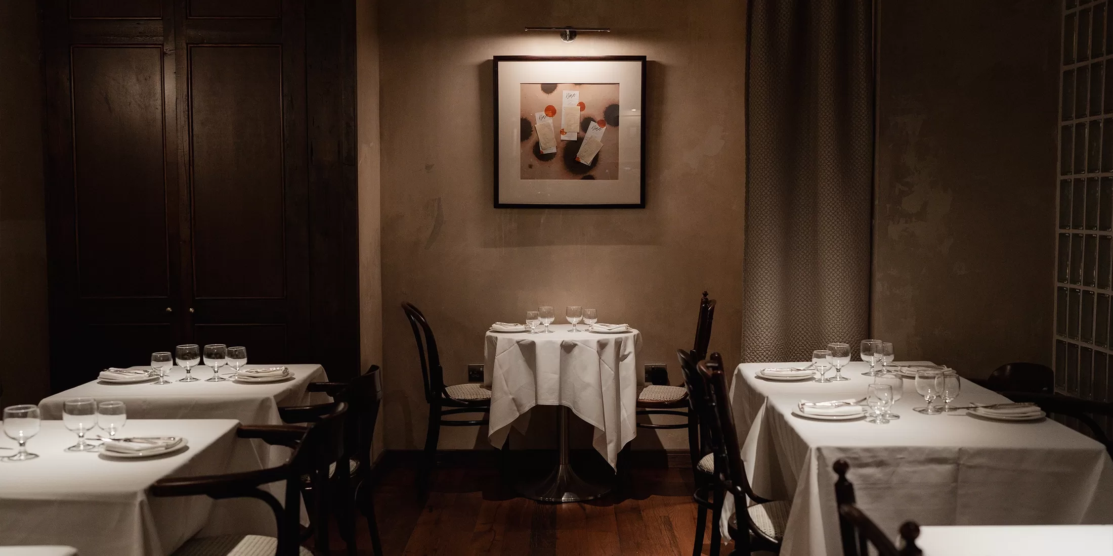 A dimly lit, elegant restaurant with white tablecloths, neatly arranged tables and chairs, and a framed abstract painting on the wall above a small table set for two.