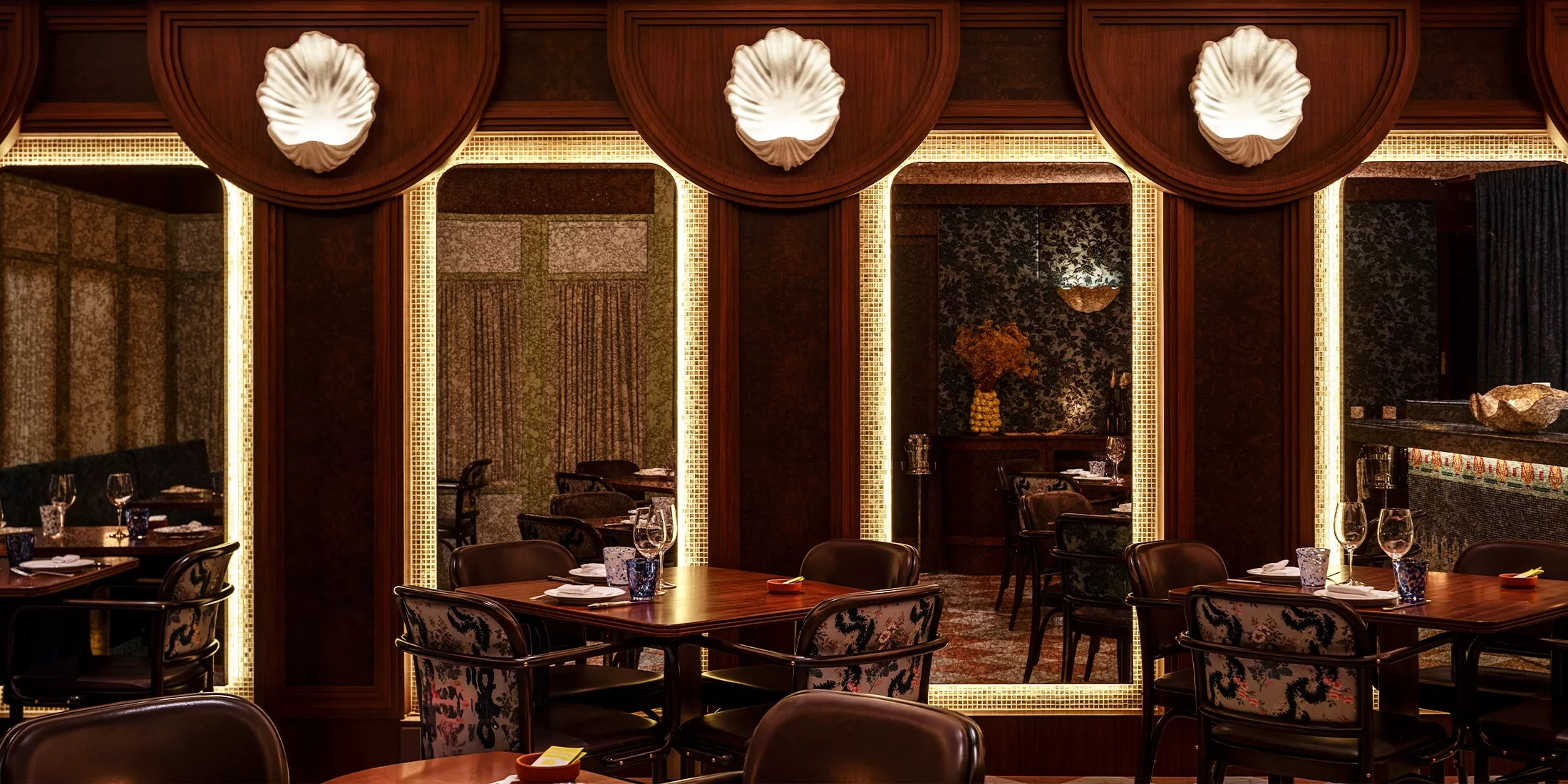 Elegant restaurant interior with dark wood walls, illuminated mirrors, and decorative shell-shaped lights above each mirror. Tables are set for dining with floral upholstery on chairs and soft, warm lighting.