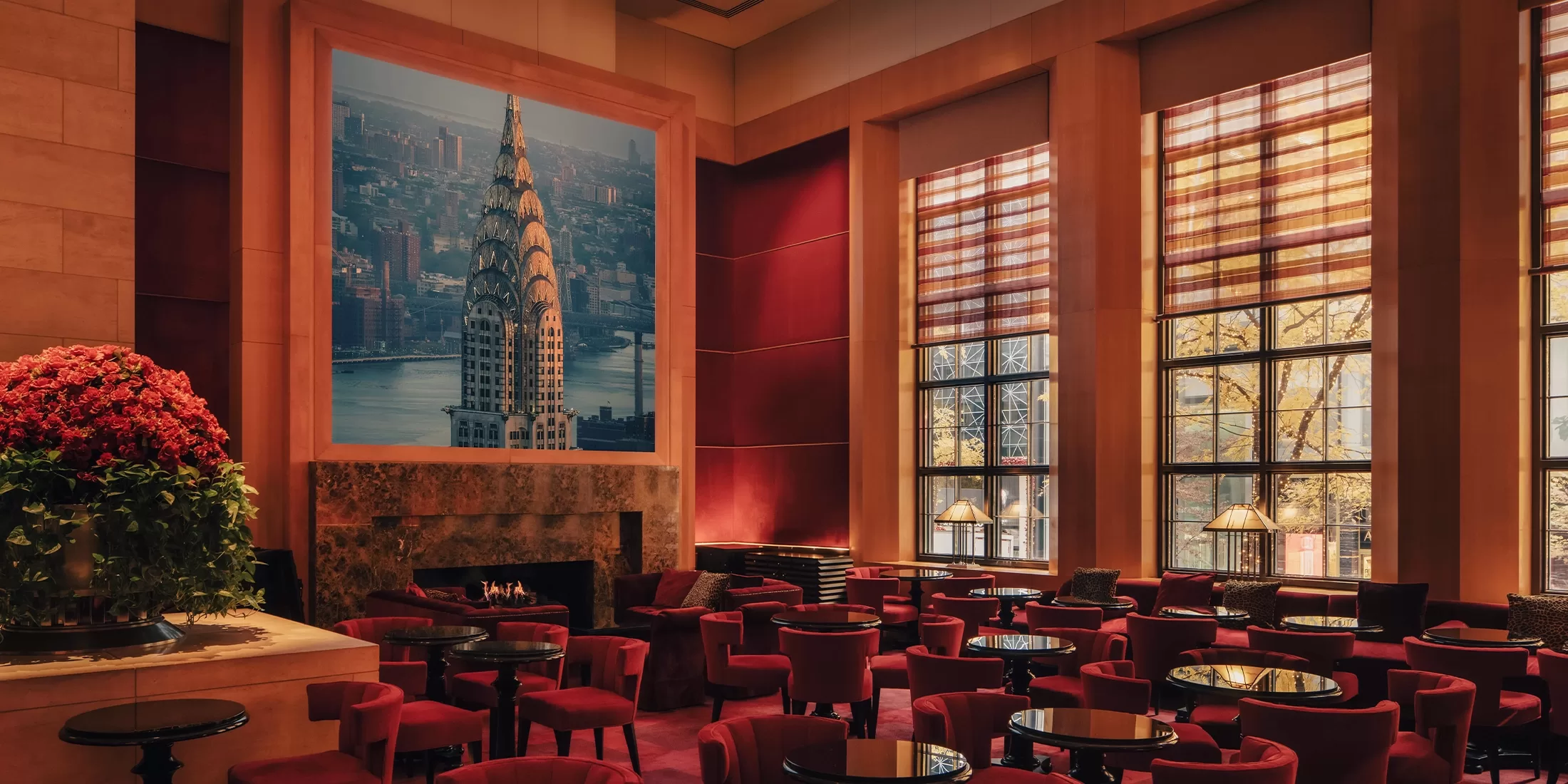 Elegant lounge with red velvet chairs and round tables, large windows with striped blinds, a fireplace, and a large framed photo of the Chrysler Building on the wall; sunlight streams through the windows.