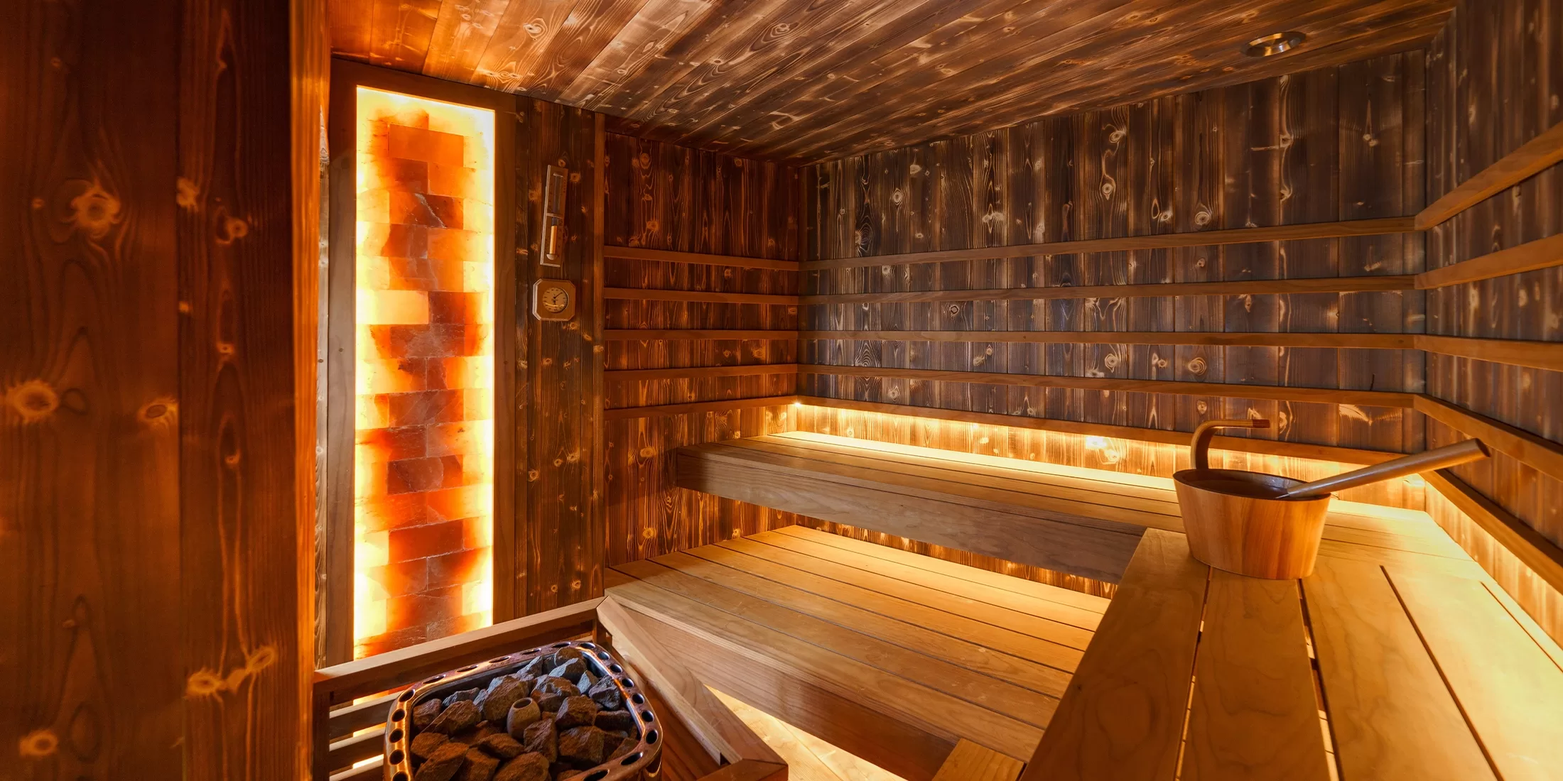 A warmly lit wooden sauna room with tiered benches, a bucket and ladle on a table, glowing backlighting behind the slats, and a basket of sauna stones in one corner.