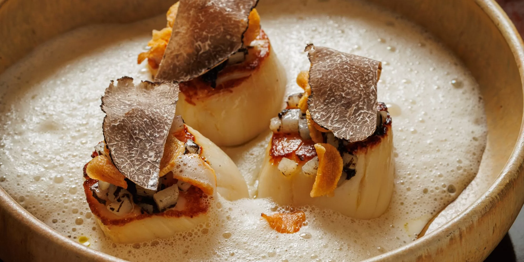 Three seared scallops topped with thin slices of black truffle and crispy garnish are served on a creamy, frothy sauce in a shallow bowl.