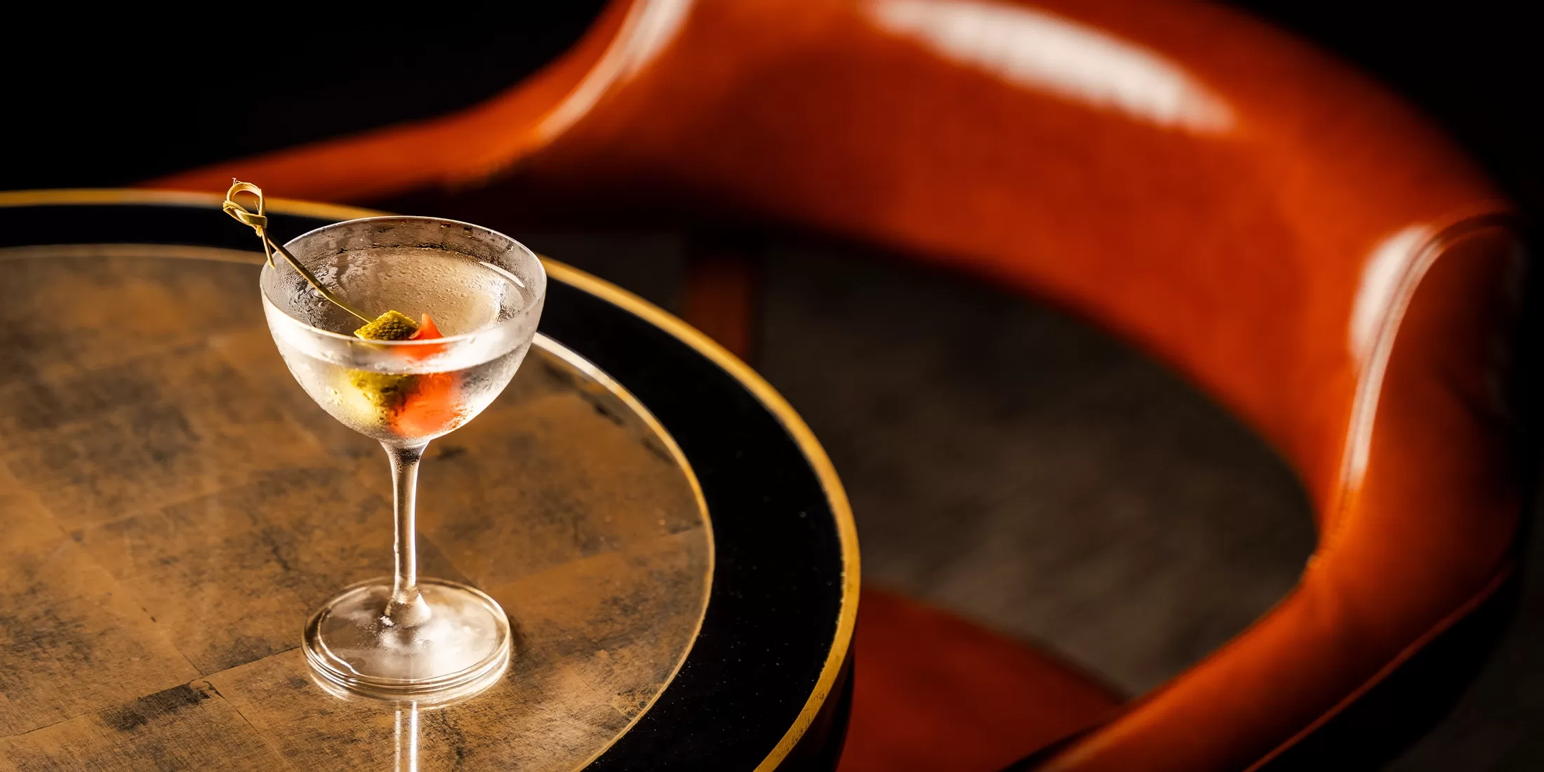 A chilled martini with olives on a cocktail pick sits on a round, gold-trimmed table next to a curved, brown leather chair in a dimly lit setting.