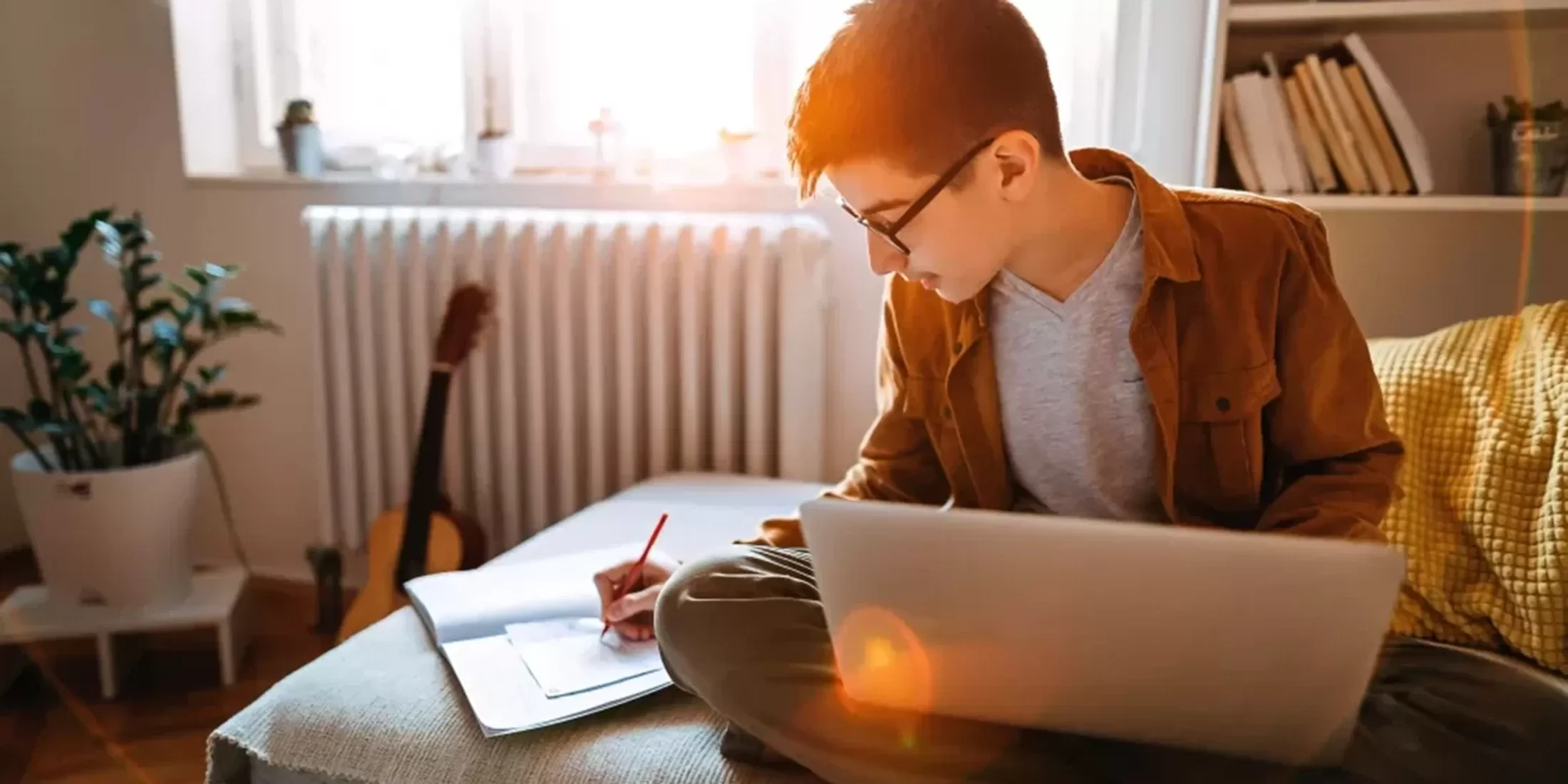 Boy with glasses crossed legged on the sofa with his laptop in his lap in a study room writing in a notepad.