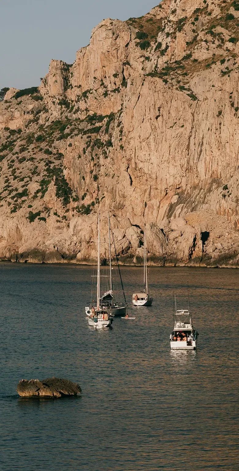 Three sailboats float on calm water near a rocky coastline with steep, rugged cliffs in the background. The sunlight casts warm tones over the scene, highlighting the natural landscape.