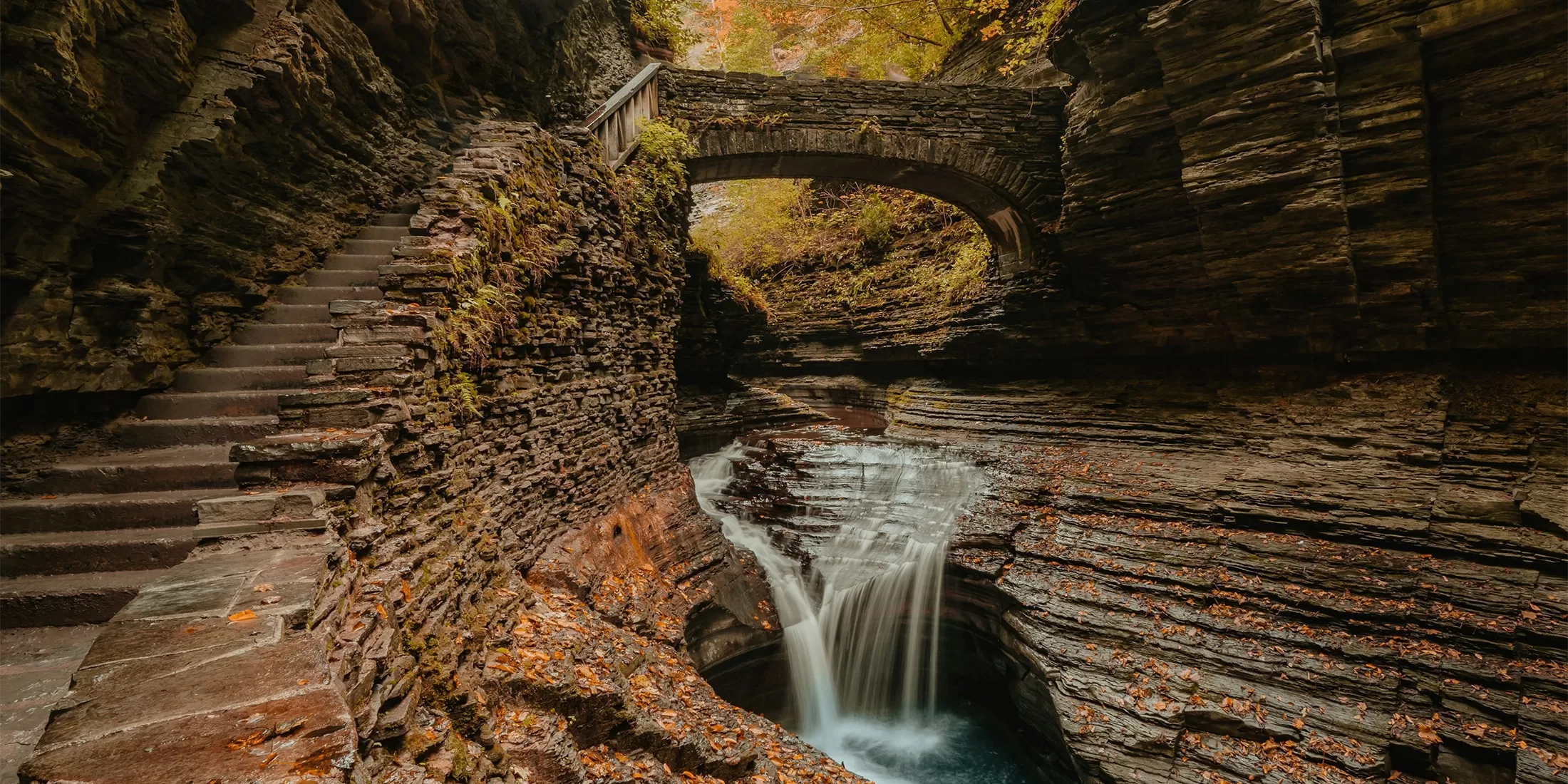 A serene waterfall cascades into a rocky gorge surrounded by autumn foliage. A stone bridge arches over the scene, and a stone staircase ascends the left side, lined with fallen leaves.