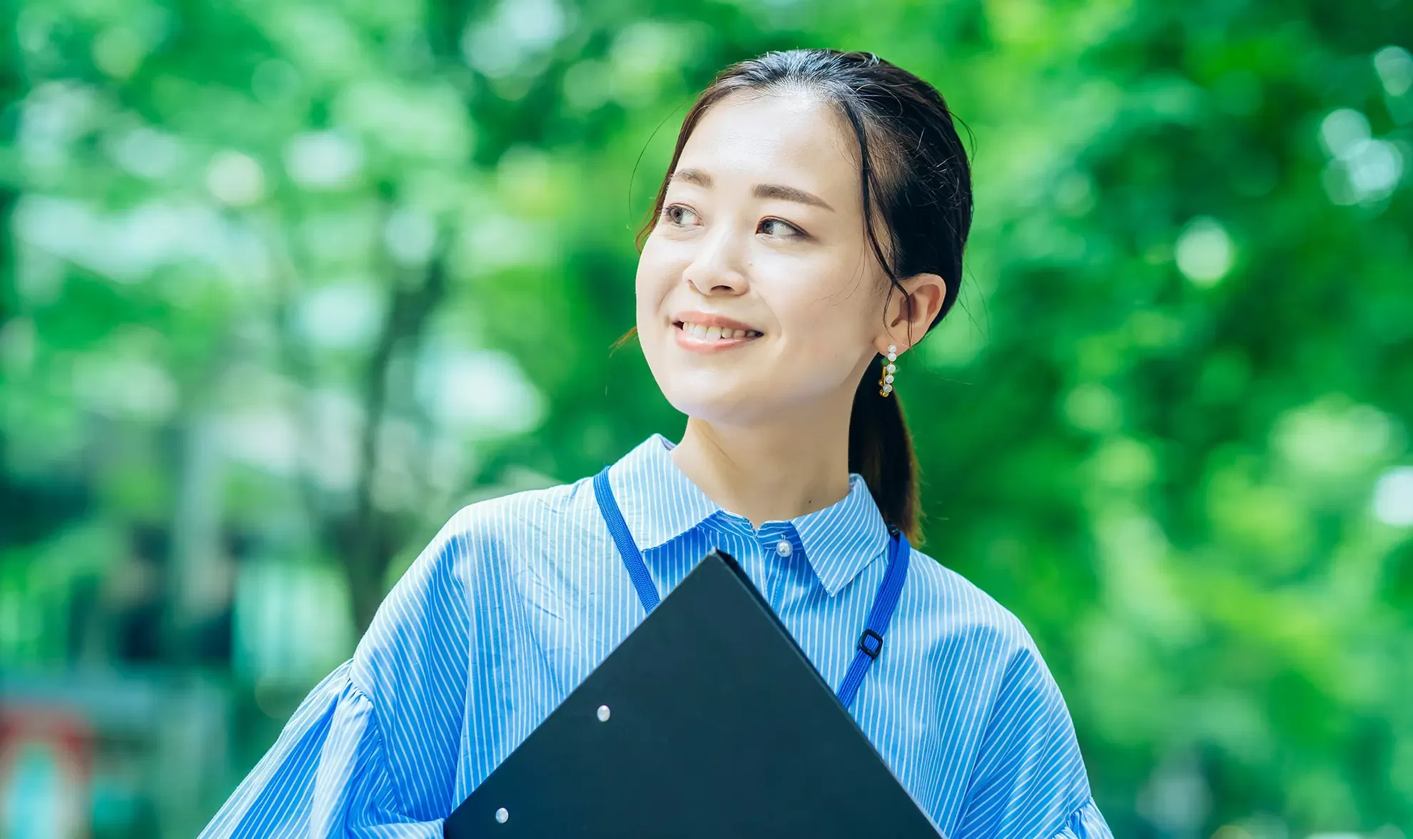A woman in a blue striped shirt smiles while holding a black file folder. She stands outdoors with green, blurred trees in the background, wearing a lanyard around her neck and gold earrings.