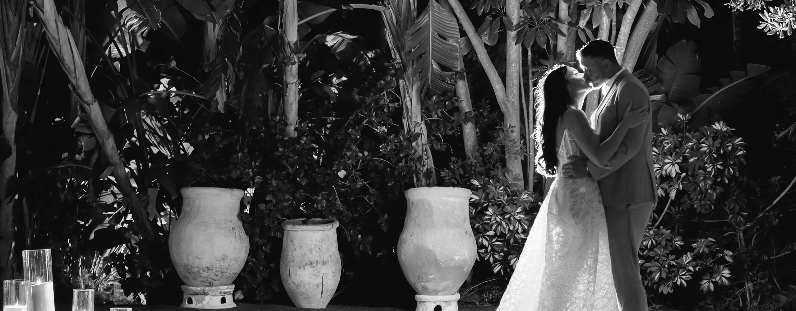 A bride and groom stand closely together at night, facing each other and smiling, surrounded by large potted plants and lush tropical foliage. The scene is lit softly, creating a romantic atmosphere.