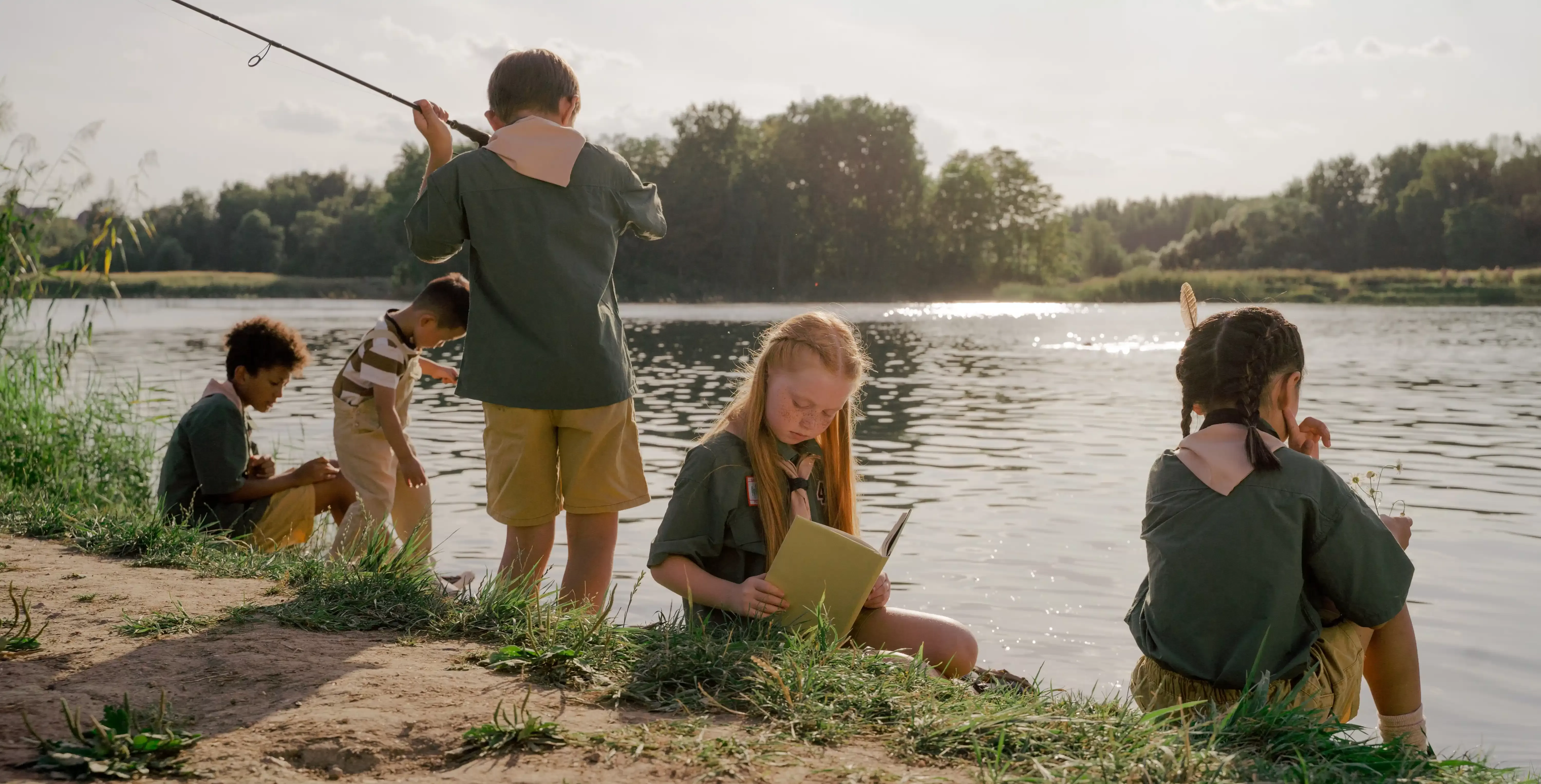 Children fishing, reading and playing by the river at summer camp