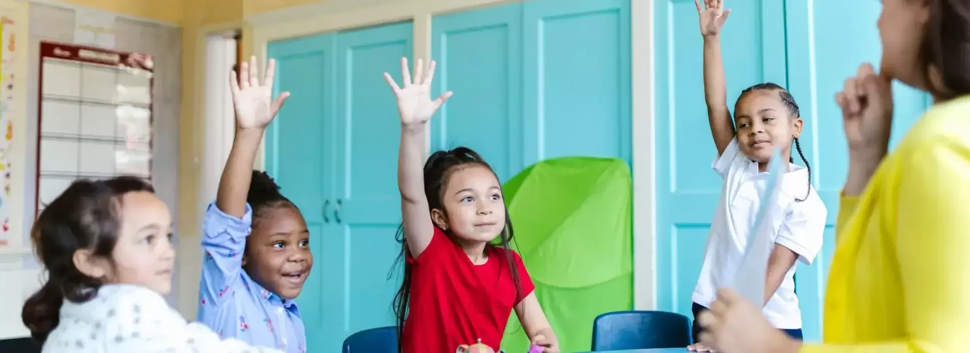 Four young children in a classroom sit at a table, three with hands raised, smiling and looking toward a teacher standing in front of them. Blue cabinets and a green board are visible in the background.
