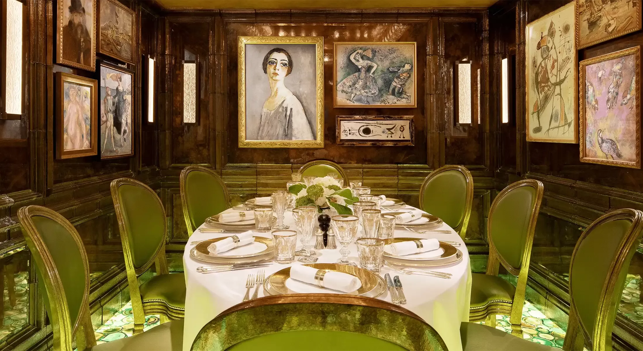 An elegant dining room with a round table set for a formal meal. The table is adorned with white linens, glassware, and floral centerpieces. Surrounding the room are wood-paneled walls decorated with framed artwork and green upholstered chairs.