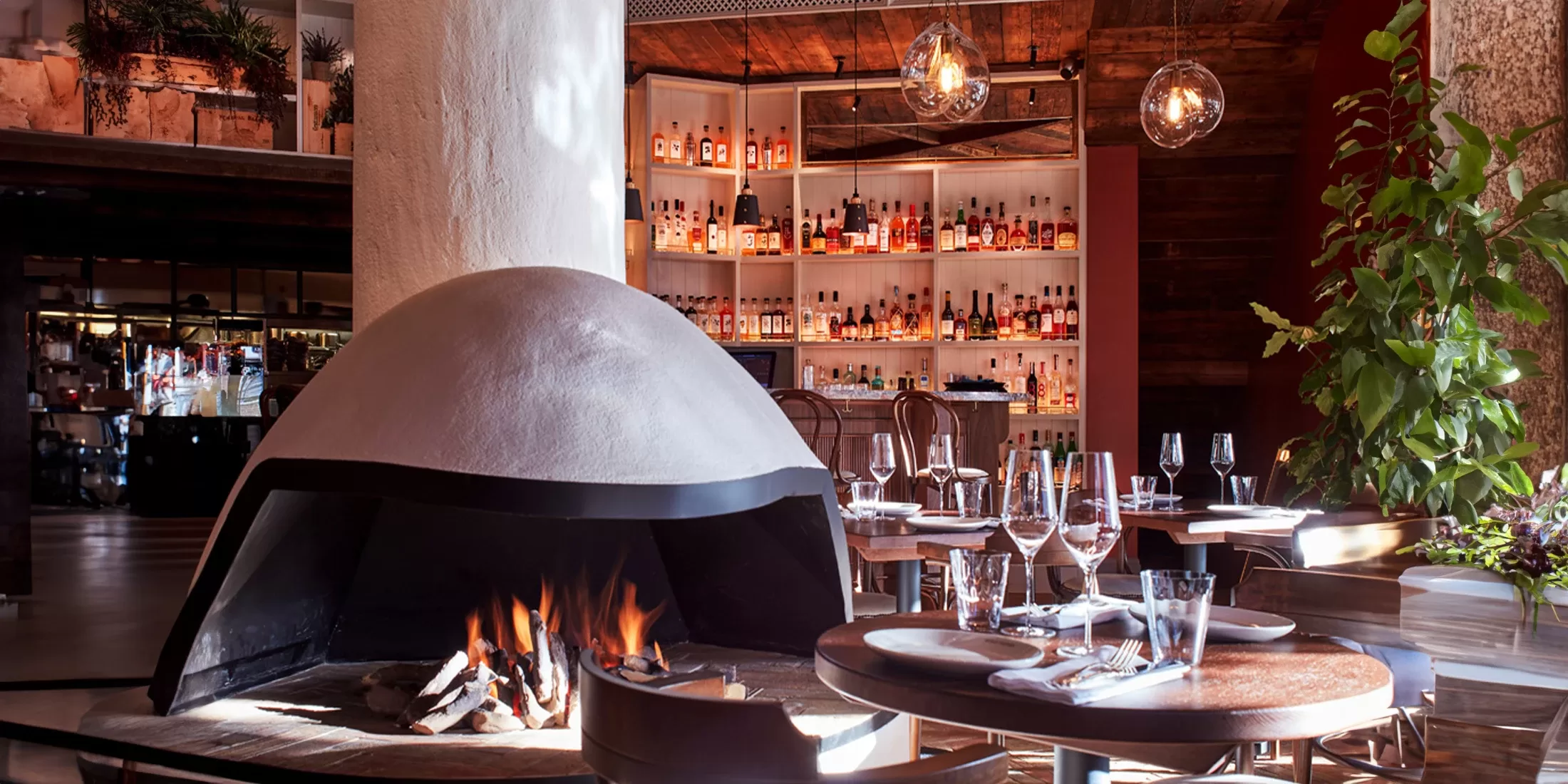 Cosy restaurant interior with a central dome-shaped fireplace, wooden tables set with glassware, and a well-stocked bar in the background. Warm lighting creates an inviting atmosphere, while plants add a touch of greenery.