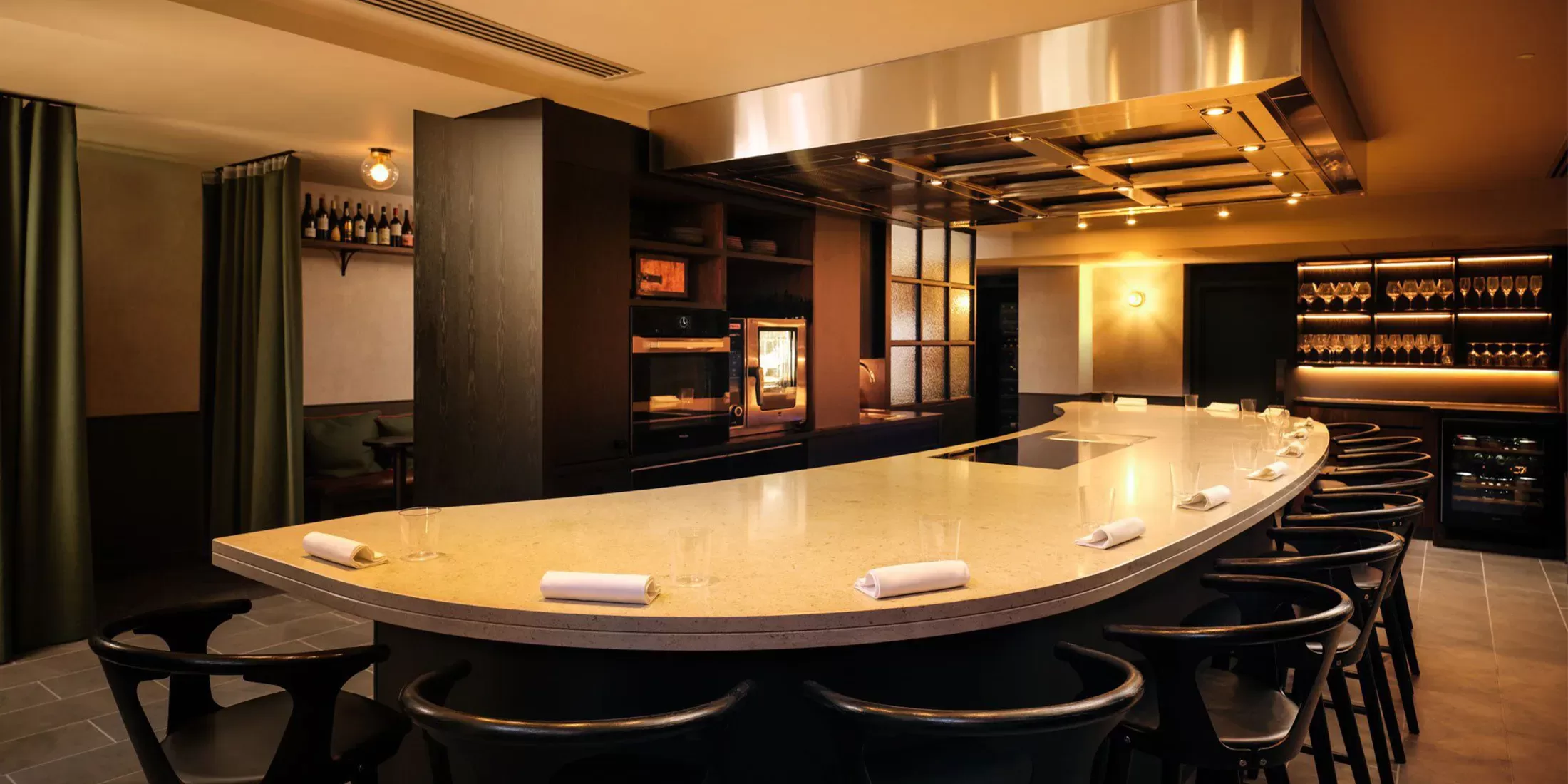 A modern, upscale restaurant interior with a curved countertop bar surrounded by sleek black chairs. Warm lighting illuminates the space, highlighting wooden shelves with bottles. White napkins are neatly placed on the bar.