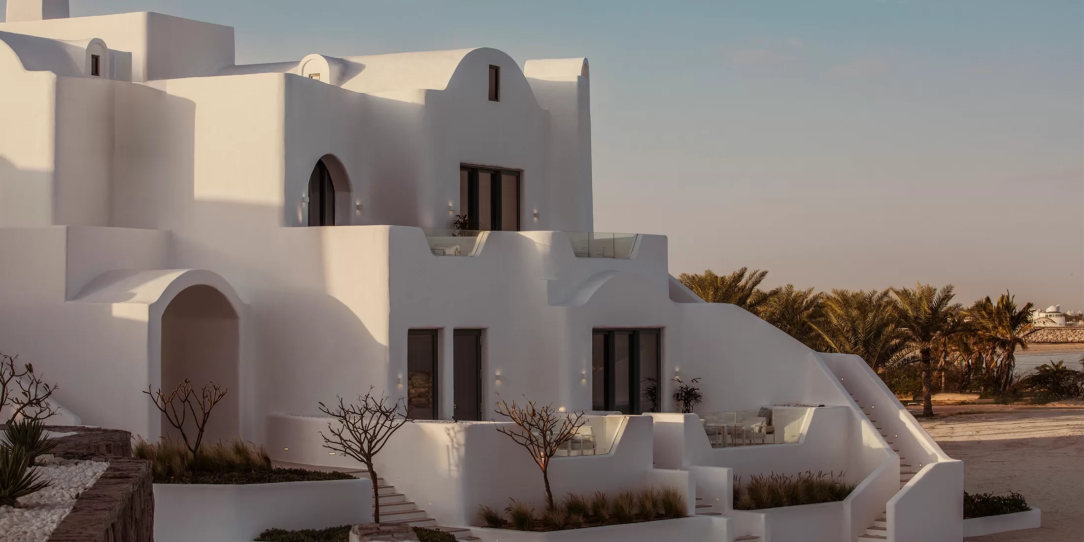 A modern white building with rounded corners stands against a clear blue sky. It features multiple levels and balconies, with surrounding greenery and a glimpse of the sea in the background. The light casts soft shadows on the structure.