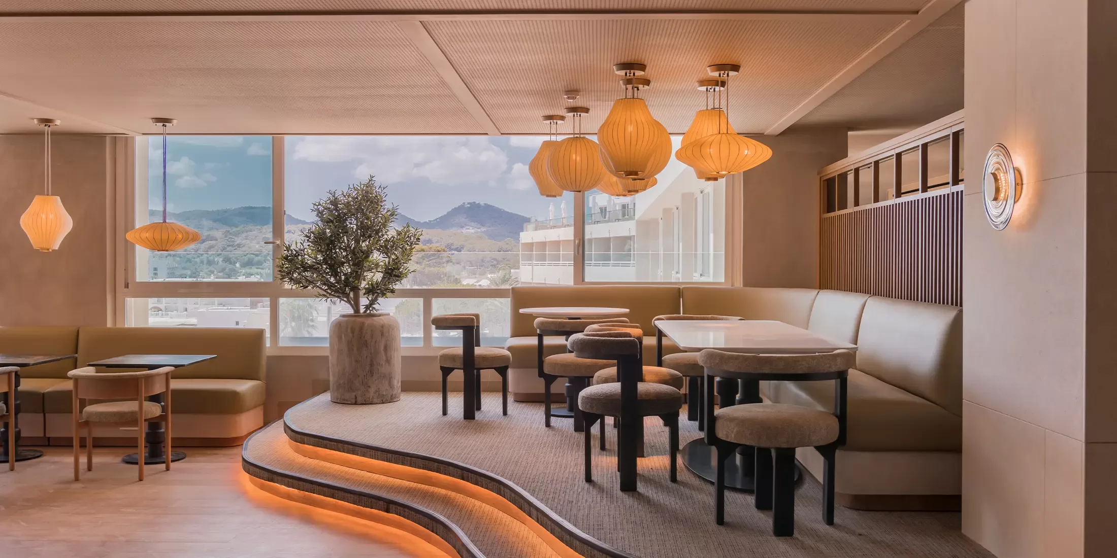 A modern cafe interior featuring stylish tables and cushioned seating on tiered levels. Soft lighting from hanging lamps creates a warm ambiance. Large windows reveal a scenic mountain view outside. A potted tree adds greenery to the space.