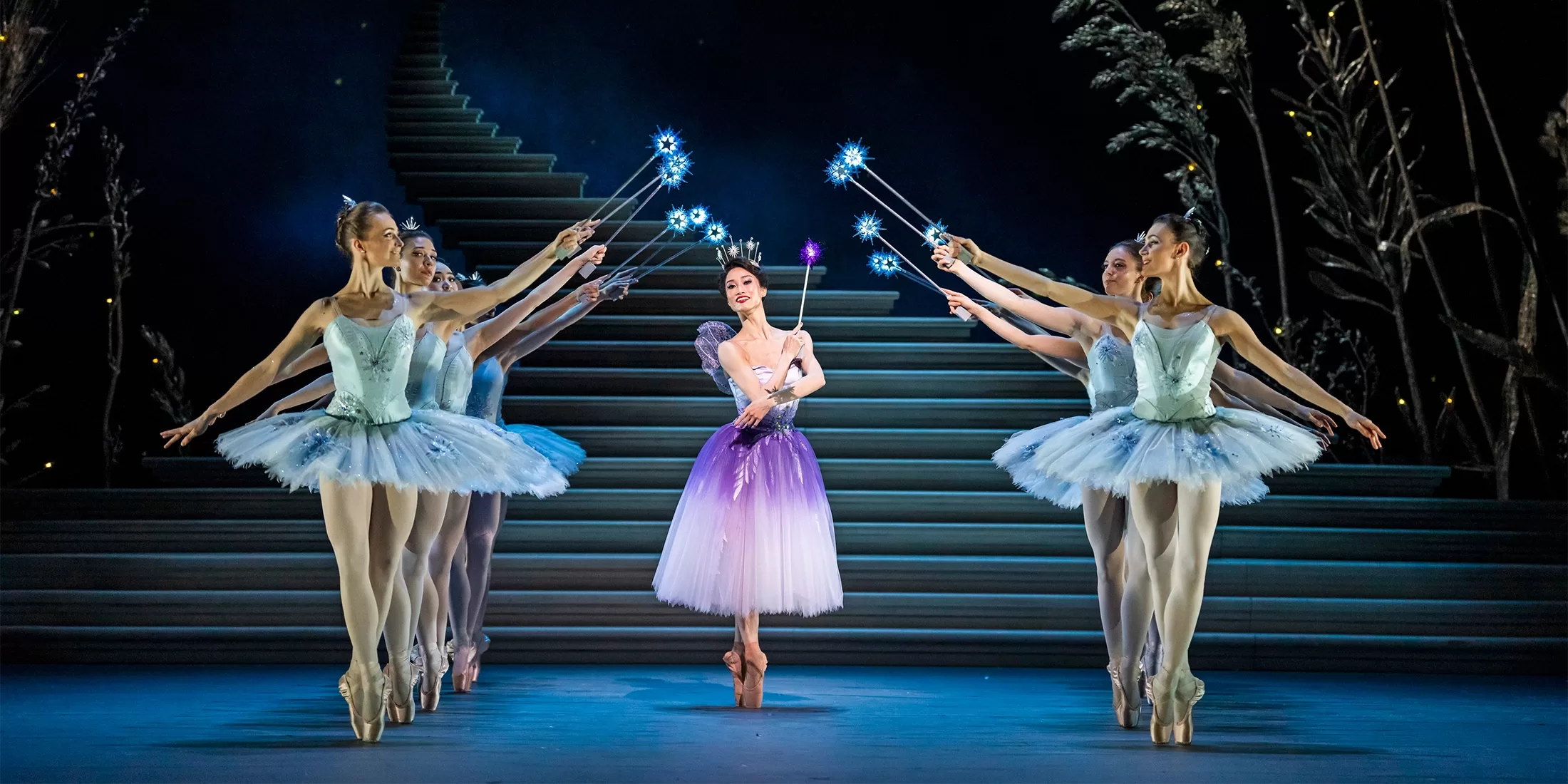 A ballerina in a purple and white tutu stands on stage, surrounded by five dancers in blue tutus. They hold sparkling wands, poised elegantly on a staircase backdrop, creating a magical, fairytale-like scene at Royal Ballet & Opera’s Cinderella.