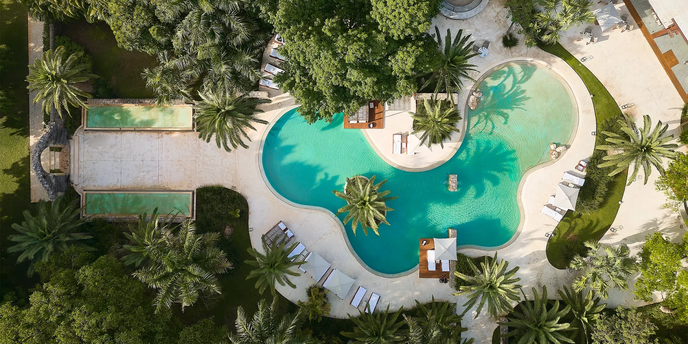 Aerial view of a landscaped outdoor pool area surrounded by lush greenery and palm trees. The pool has a curvy shape with lounge chairs and umbrellas around it. A smaller rectangular pool is adjacent on the left, with pathways connecting the spaces.