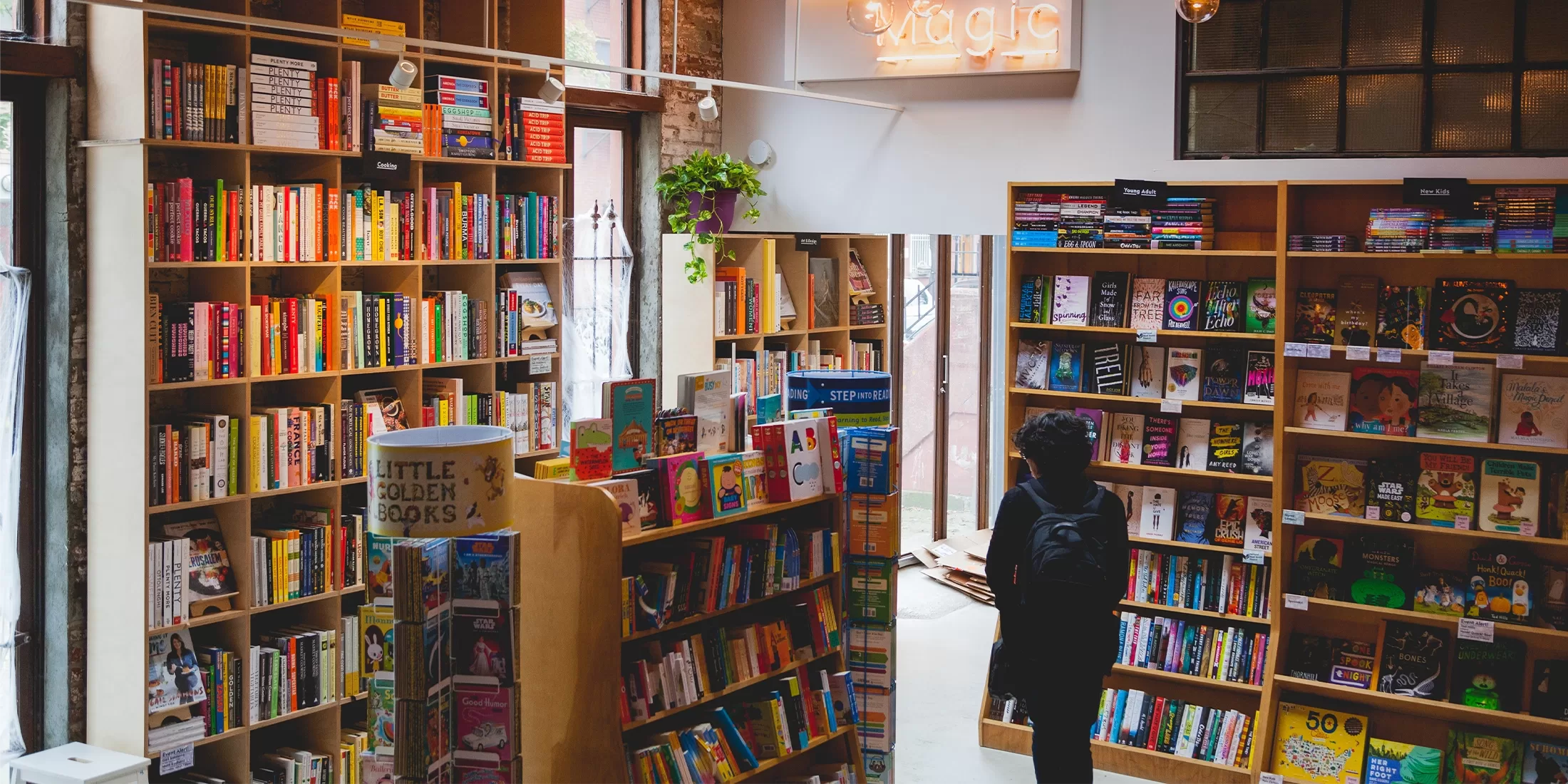 A person stands in a cosy bookstore, surrounded by tall wooden shelves filled with colorful books. A neon sign reading magic hangs on the wall. Soft light filters through large windows, creating a warm and inviting atmosphere.
